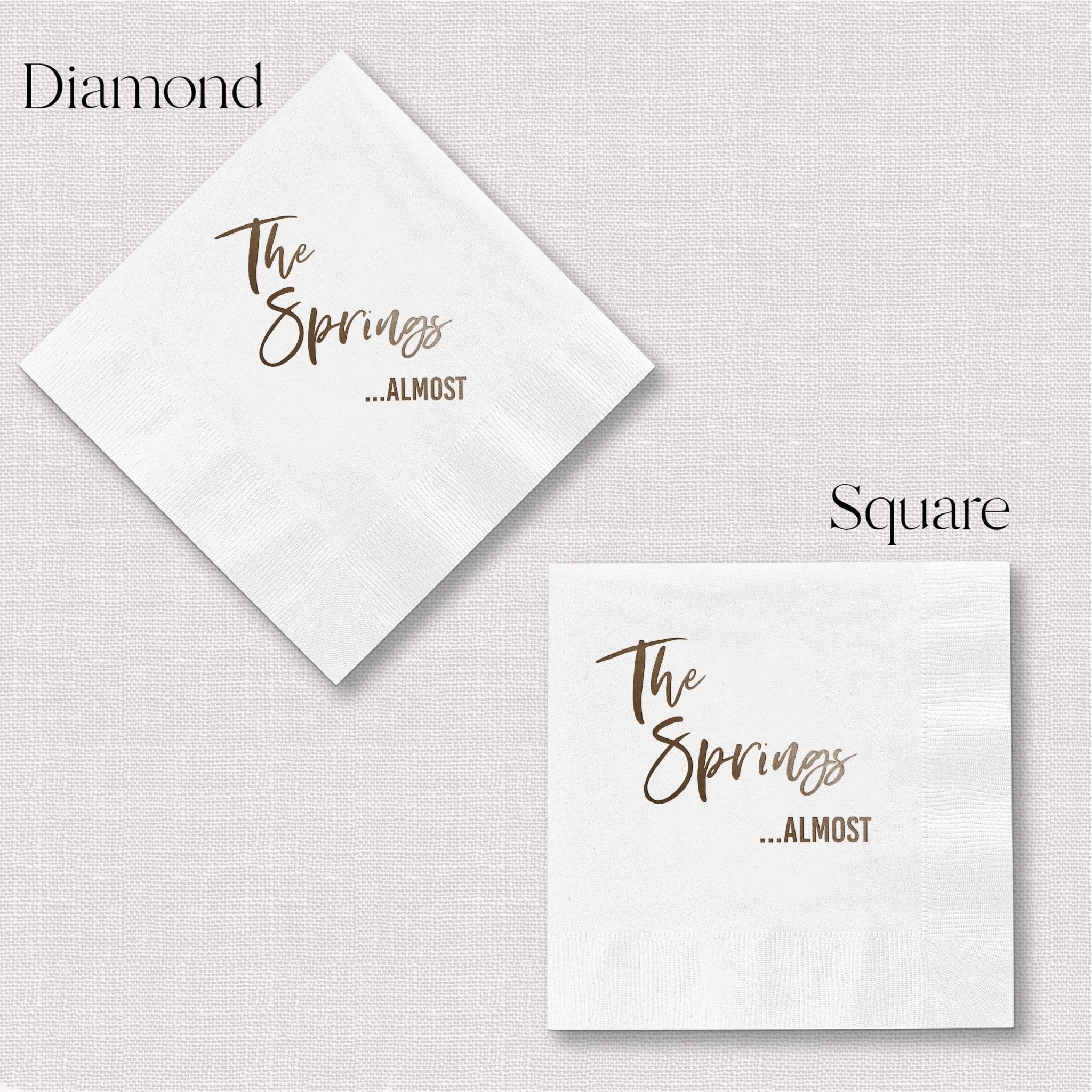 White foil stamped cocktail napkins in diamond and square shapes with "The Springs ...ALMOST" text in gold lettering.