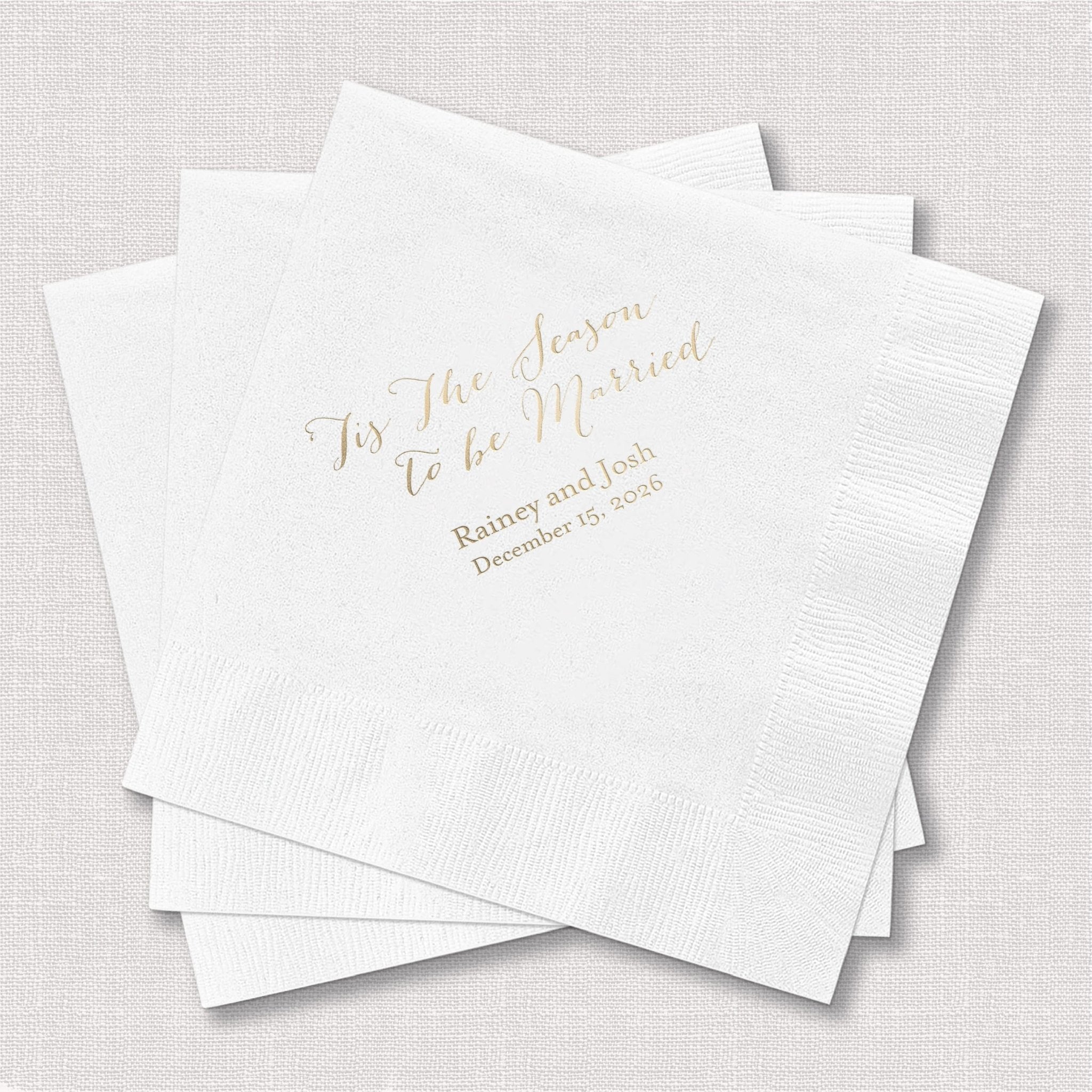 White napkins with gold foil stamped text reading "Tis The Season to be Married" and personalized names and date below.