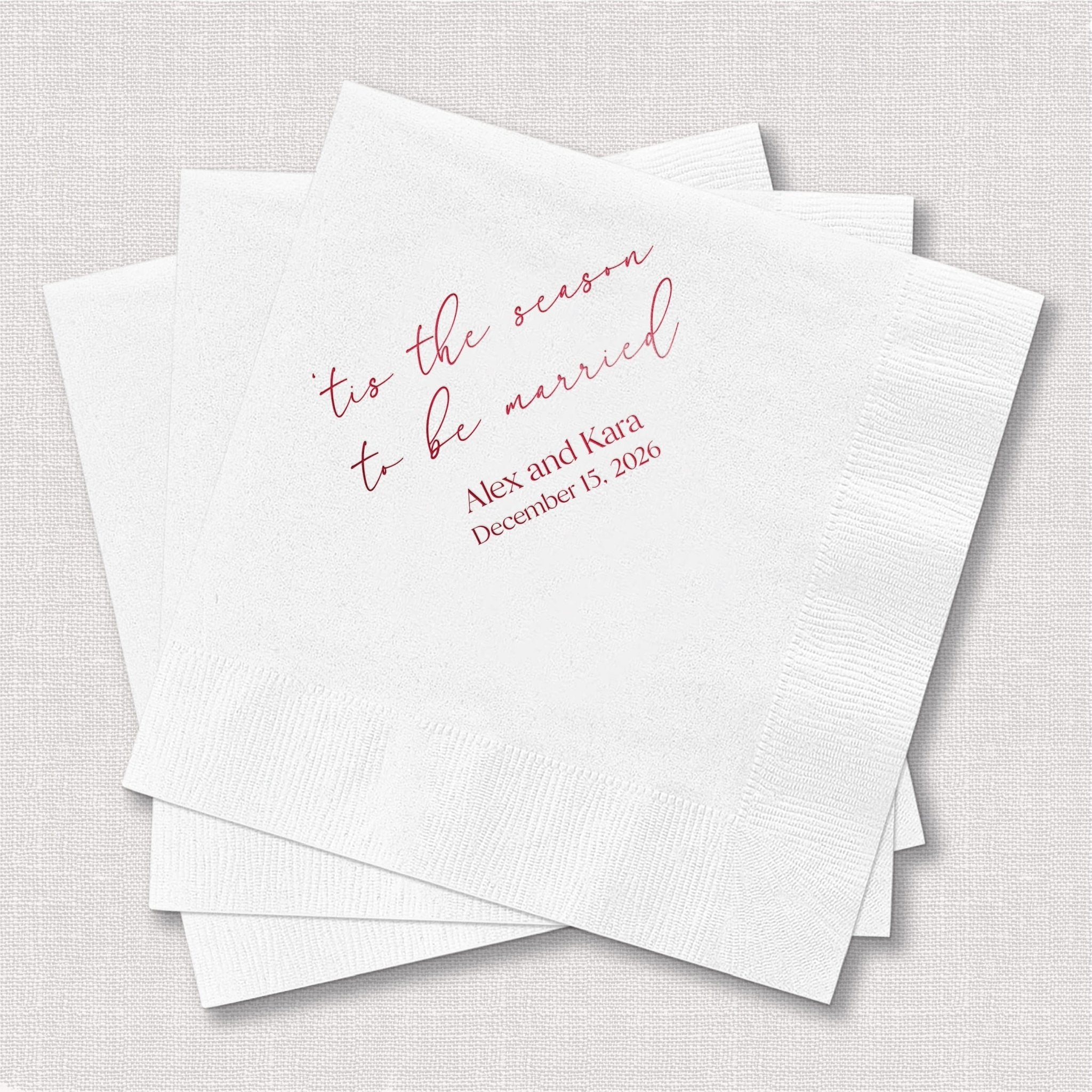 Stack of white napkins with pink foil-stamped text reading "tis the season to be married" and personalized names and date below.