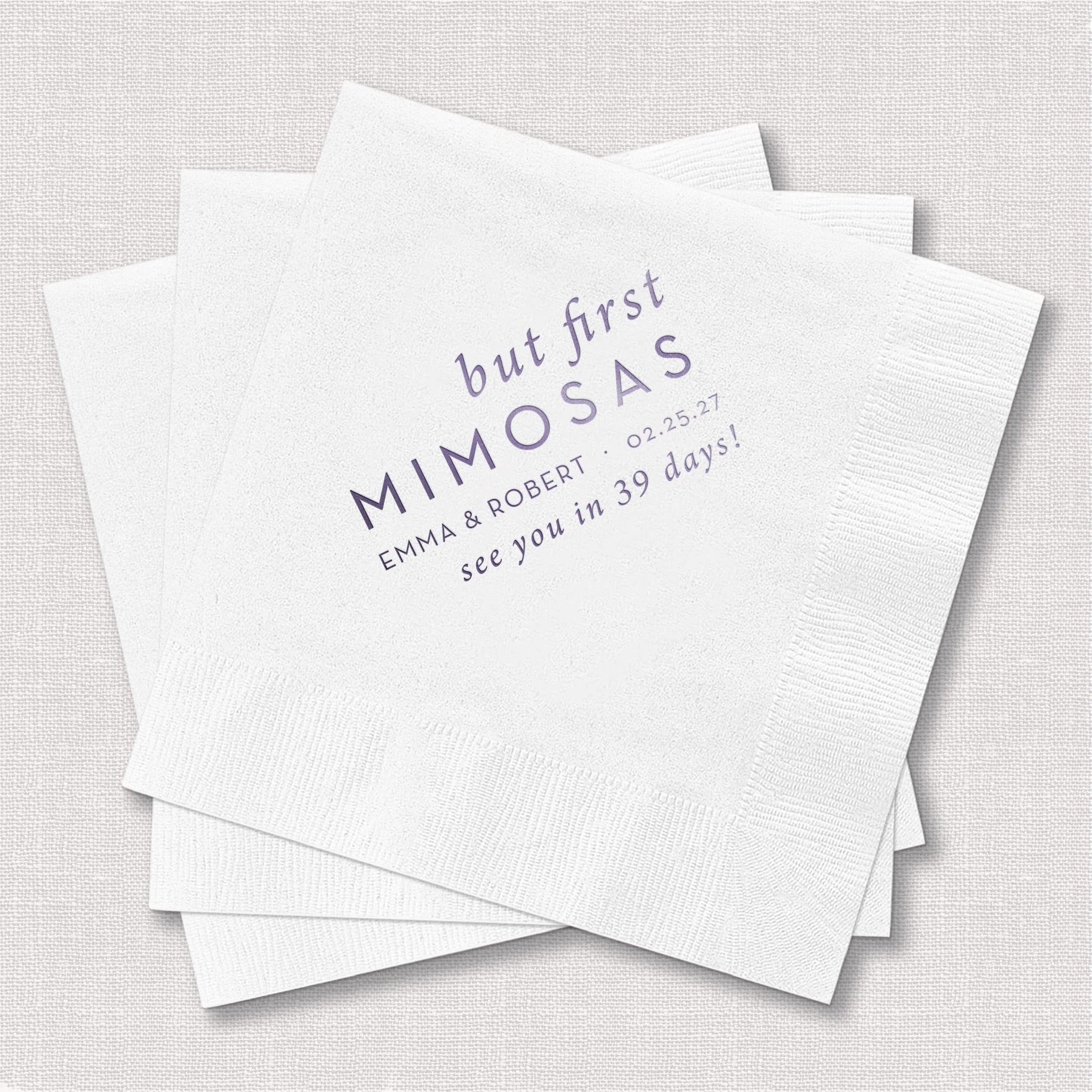 White cocktail napkins with "but first MIMOSAS" text and personalized names and date in gray foil stamping.