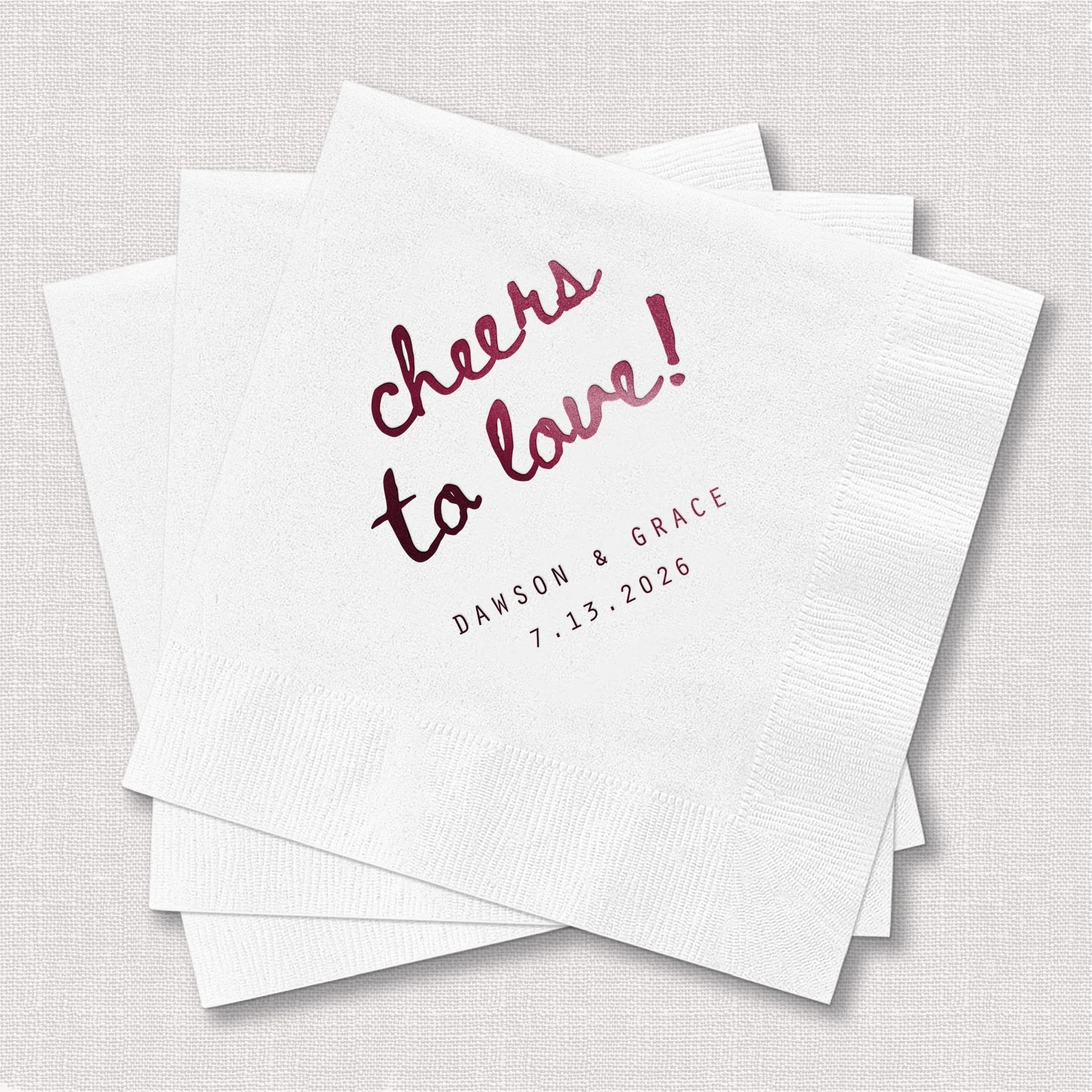 White cocktail napkins with burgundy foil-stamped "cheers to love" text and personalized names and date below.