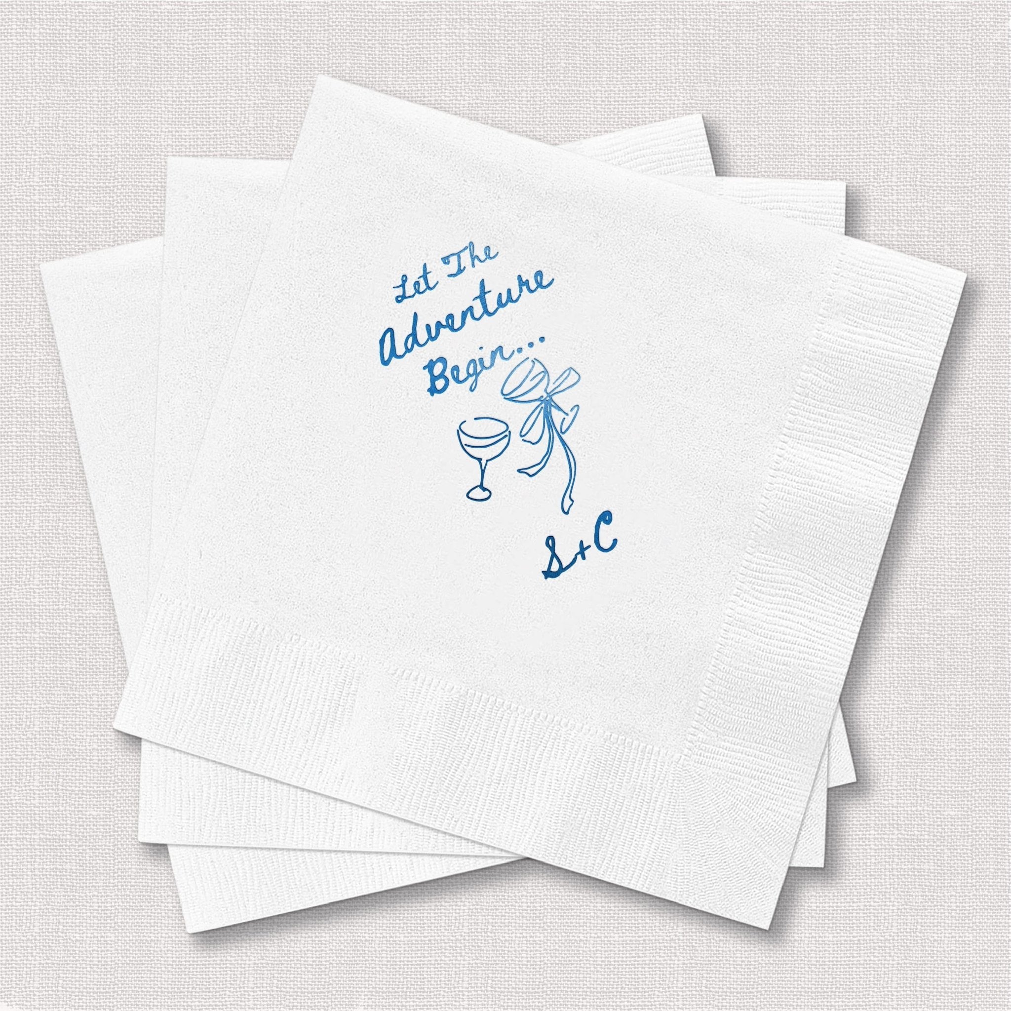Stack of white cocktail napkins with blue foil-stamped text reading "Let the Adventure Begin" and champagne glass design.