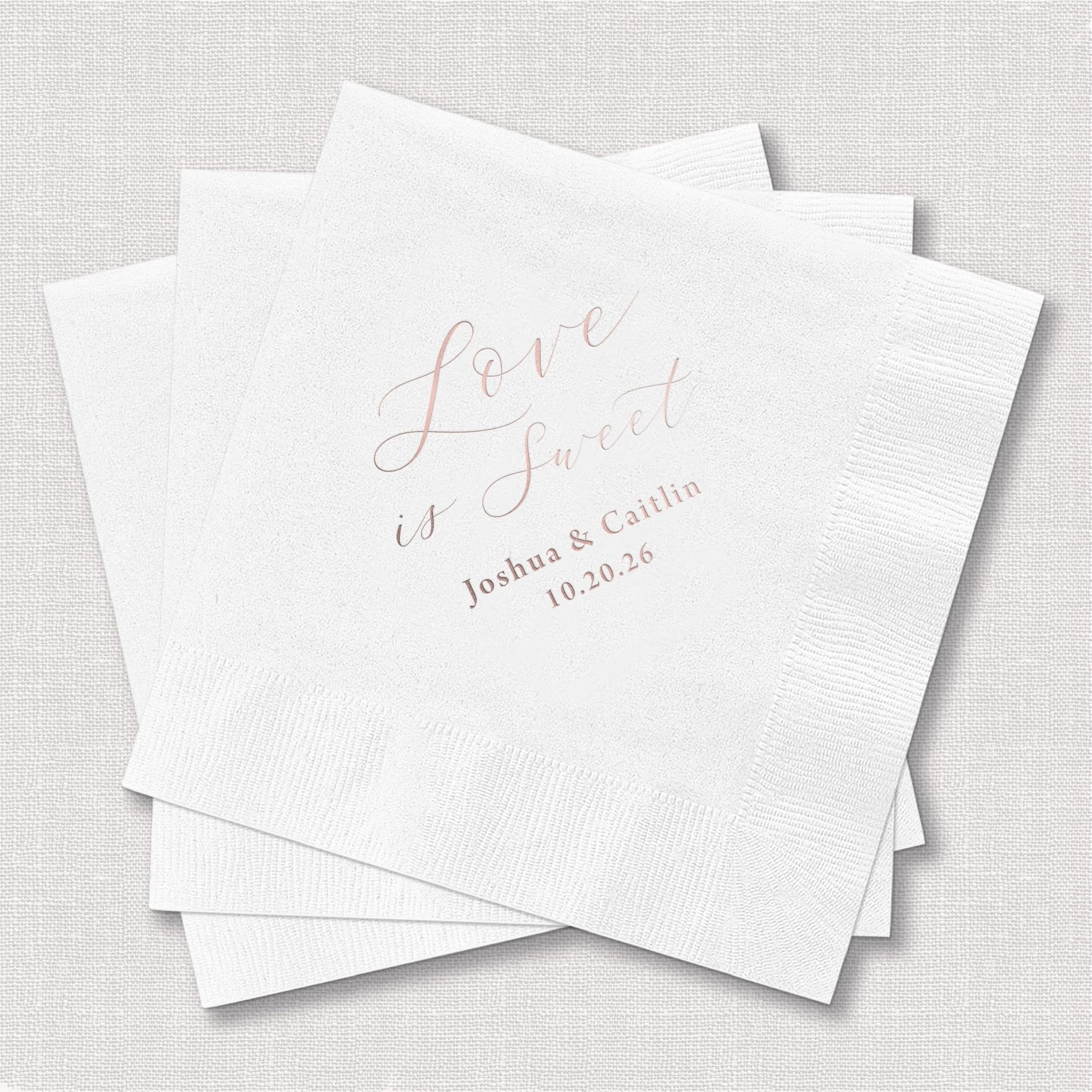 Stack of white cocktail napkins with rose gold foil stamped text reading "Love is Sweet" and personalized names and date.