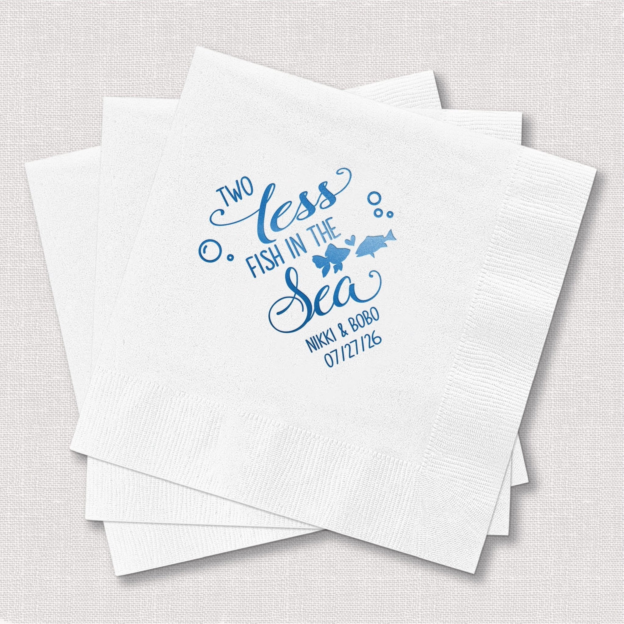 Stack of white napkins with blue foil-stamped design reading "Two Less Fish in the Sea" and personalized wedding date and couple names.