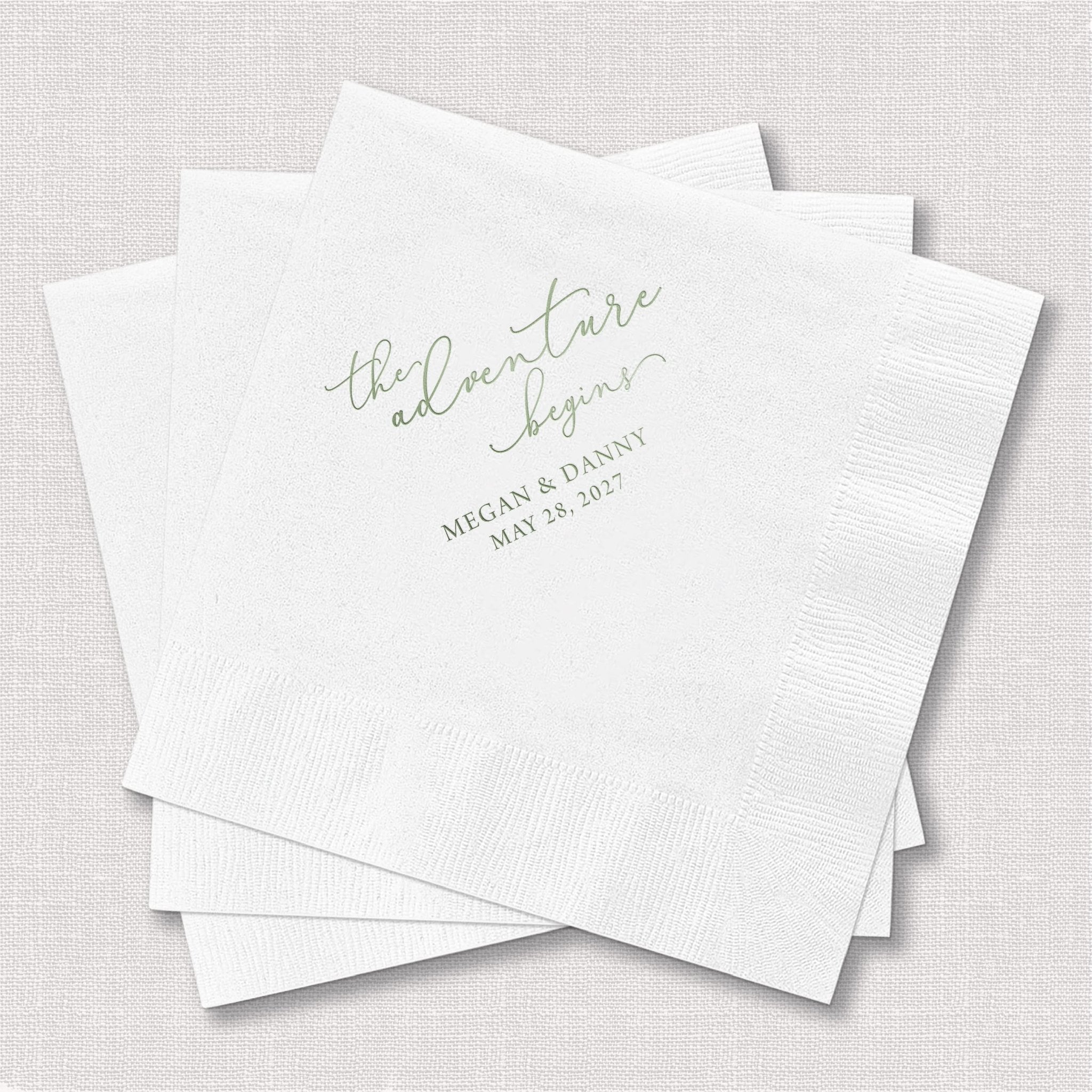Stack of white napkins with "the adventure begins" in foil lettering and personalized names and date below.