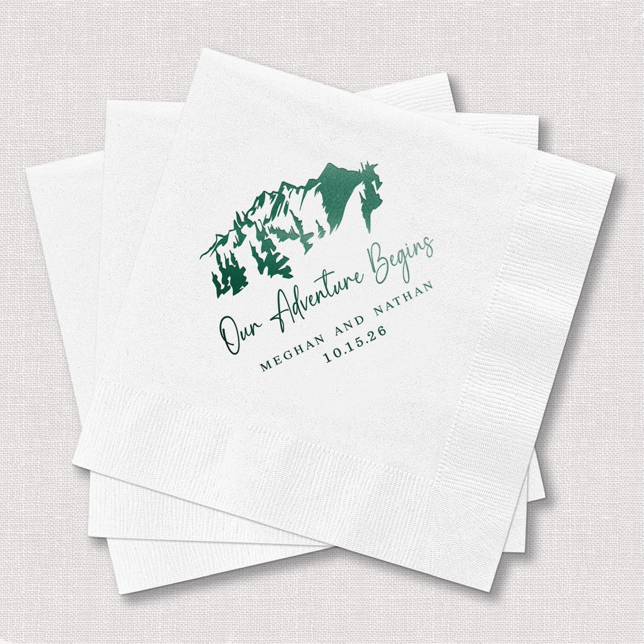 White cocktail napkins with green foil-stamped mountain design and "Our Adventure Begins" text with personalized names and wedding date.