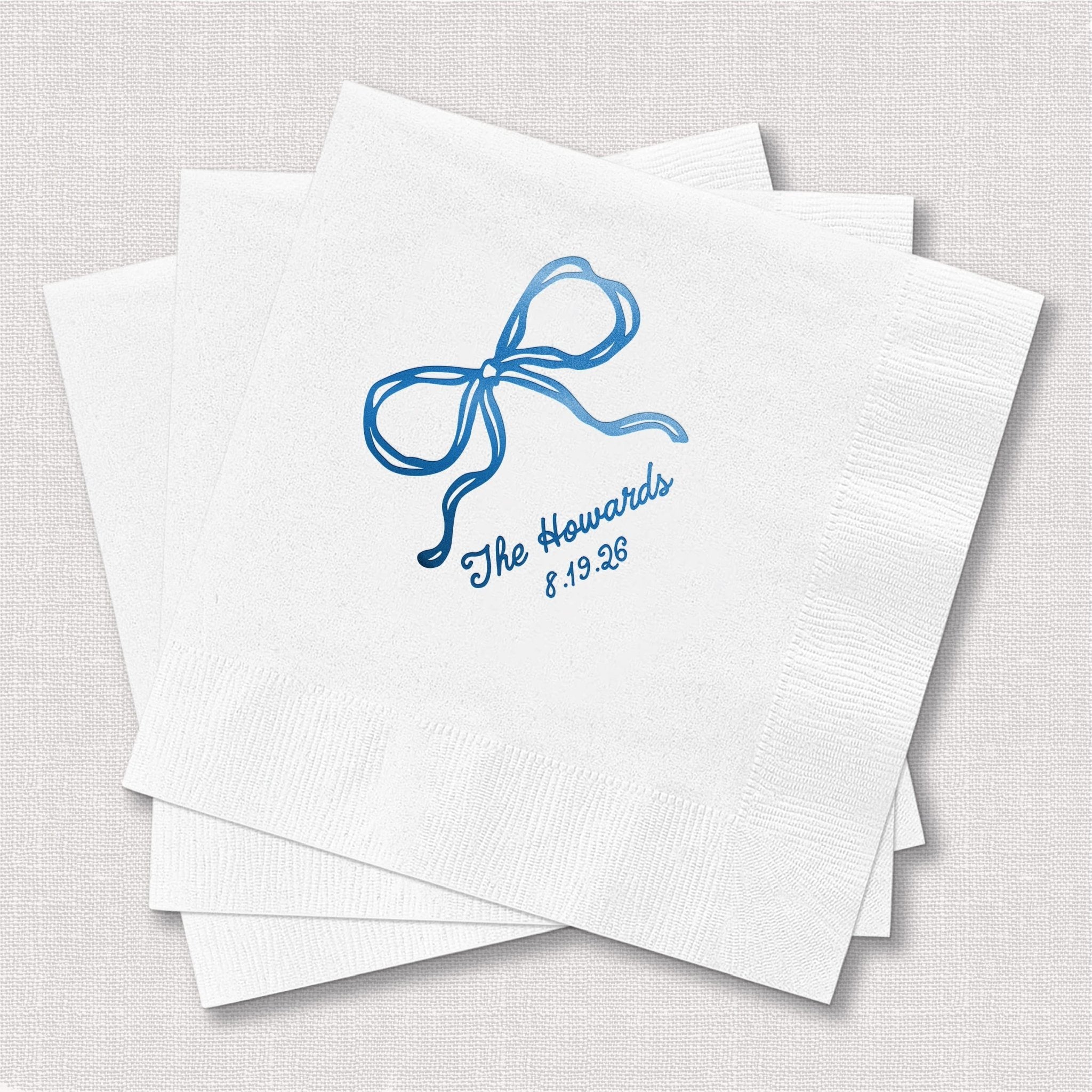 White cocktail napkins with blue foil-stamped bow design and personalized text reading "The Hayshids 5.19.23"