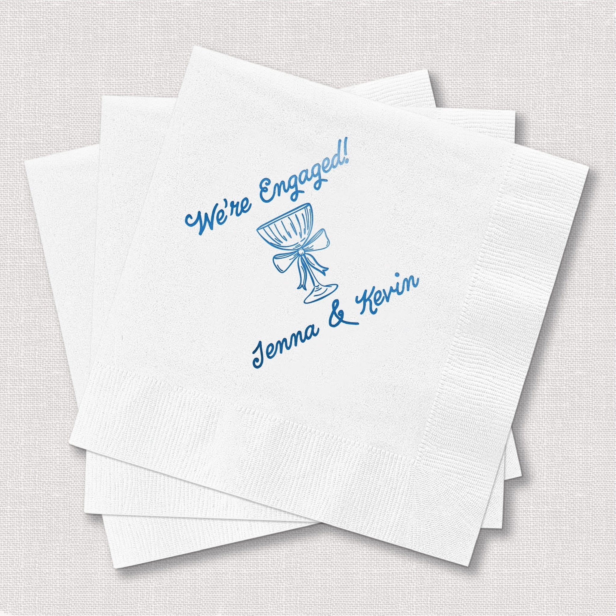 Stack of white cocktail napkins with blue foil-stamped design reading "We're Engaged" and personalized names "Jenna & Kevin" with bow illustration.