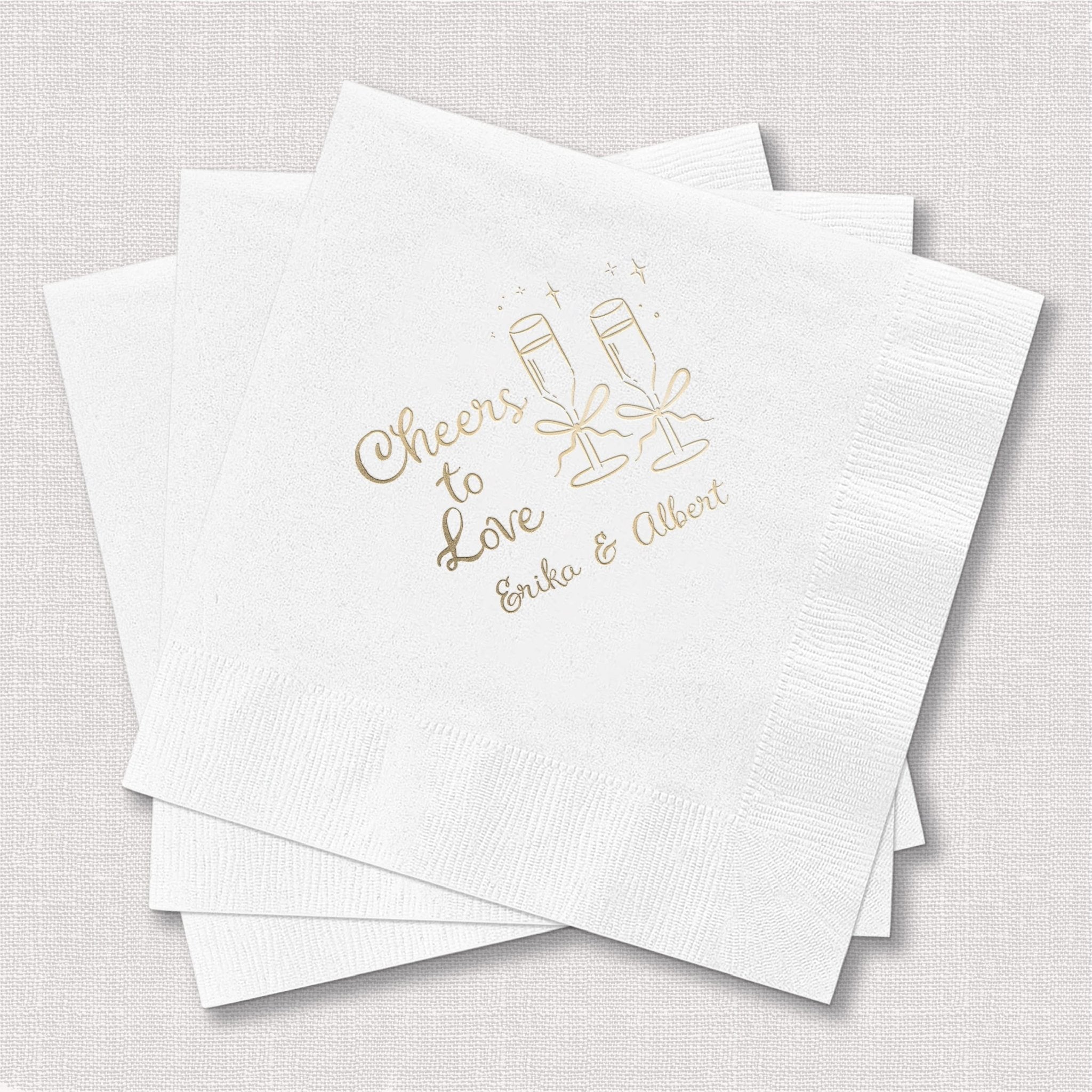 White cocktail napkins with gold foil stamped champagne glasses and "Cheers to Love" text with personalized names Erika and Allison