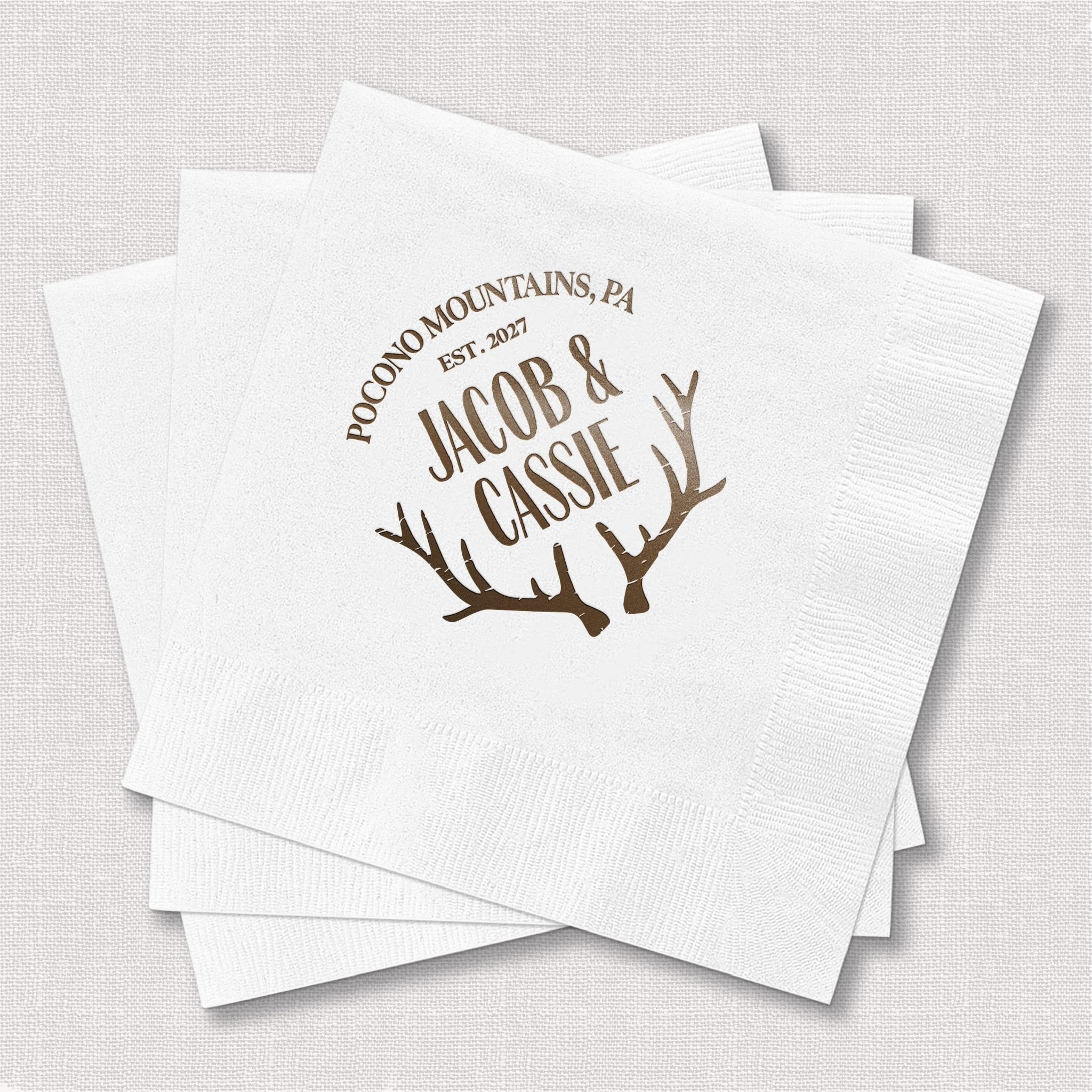 White napkins with brown foil-stamped antlers and text reading "Pocono Mountains, PA Jacob & Cassie June 2027"