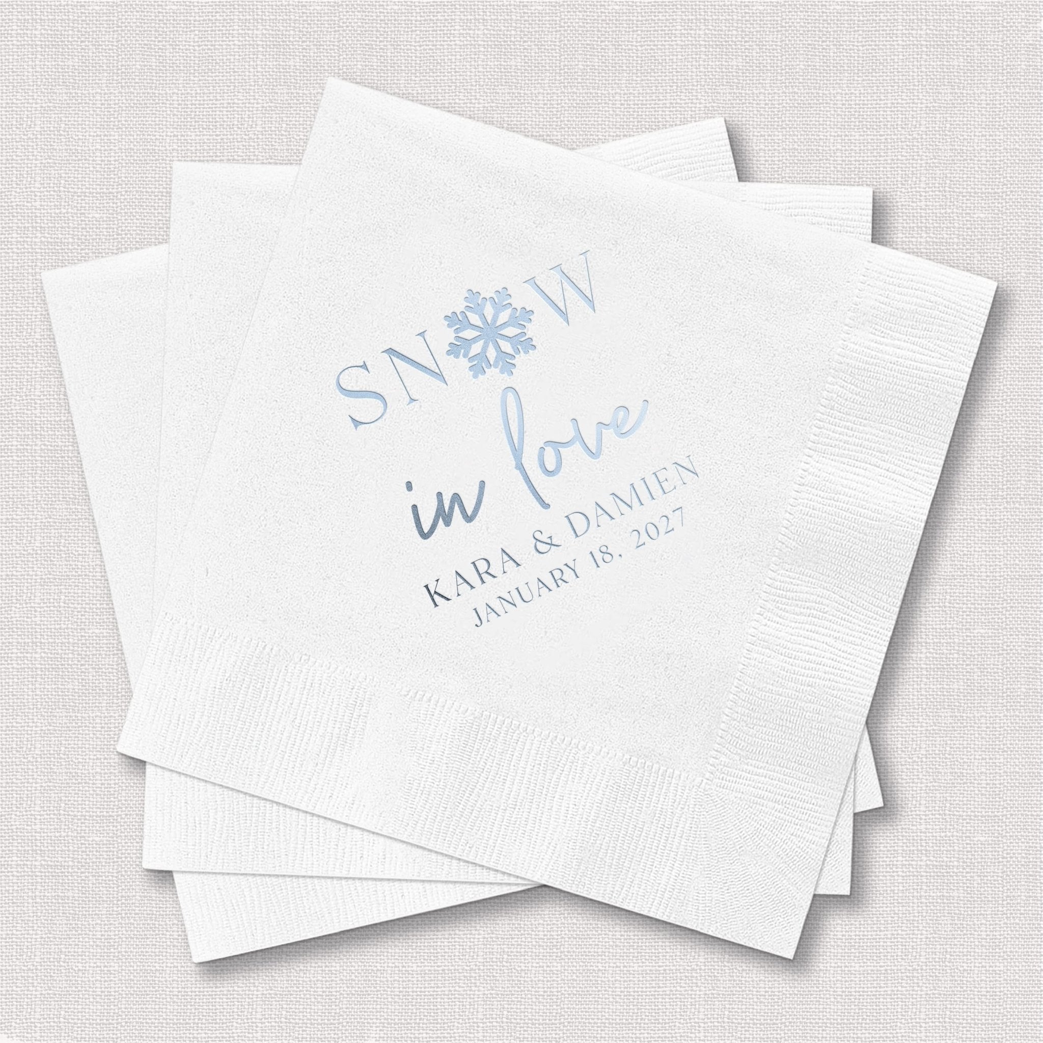 White cocktail napkins with blue foil-stamped "Snow in Love" design, names, and wedding date personalization.