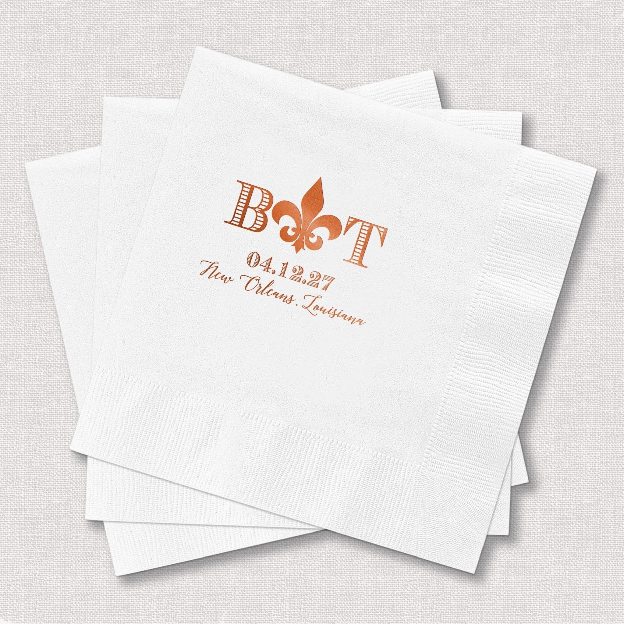 White paper napkins with orange foil-stamped fleur de lis, initials, date, and couple's names for wedding favors.