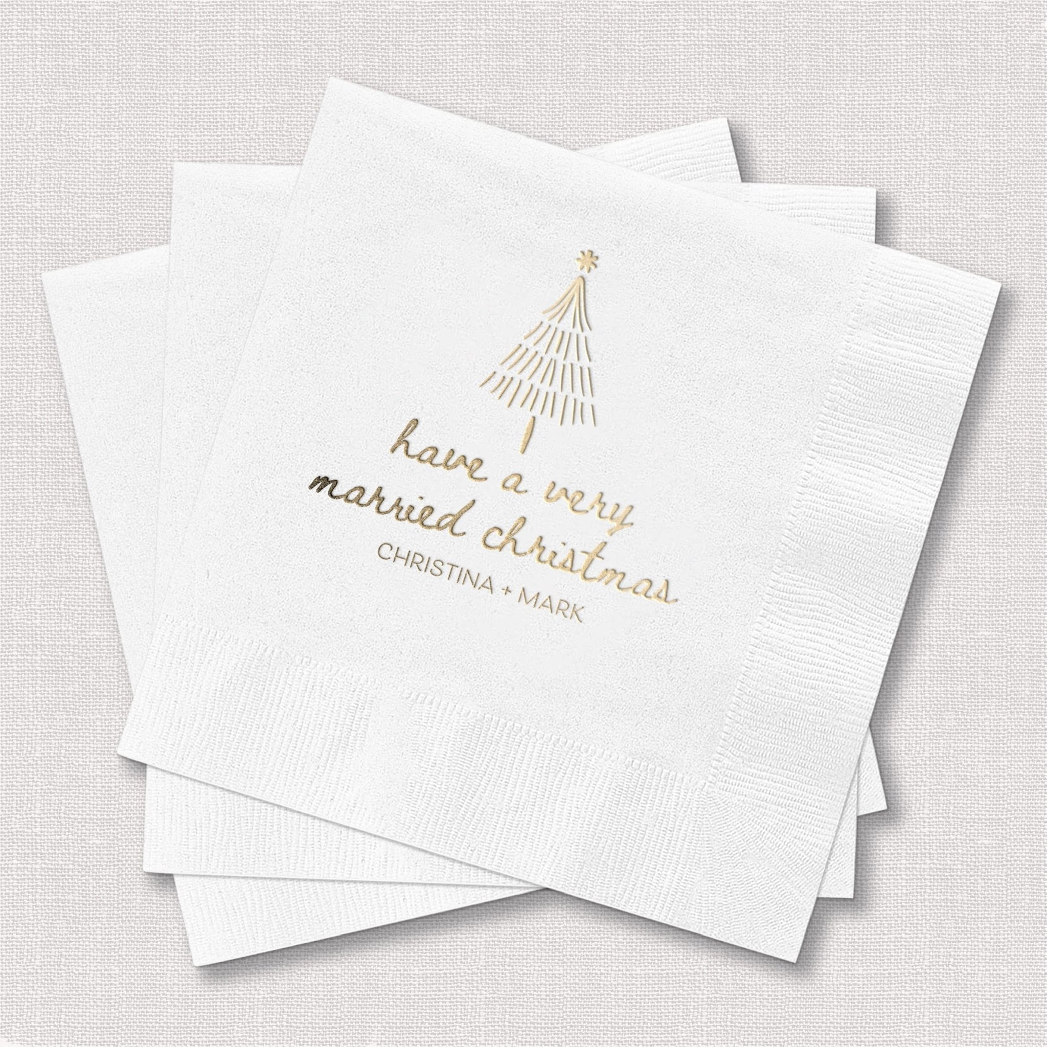 White cocktail napkins with gold foil stamped Christmas tree and personalized text reading "have a very married christmas" with couple's names below.