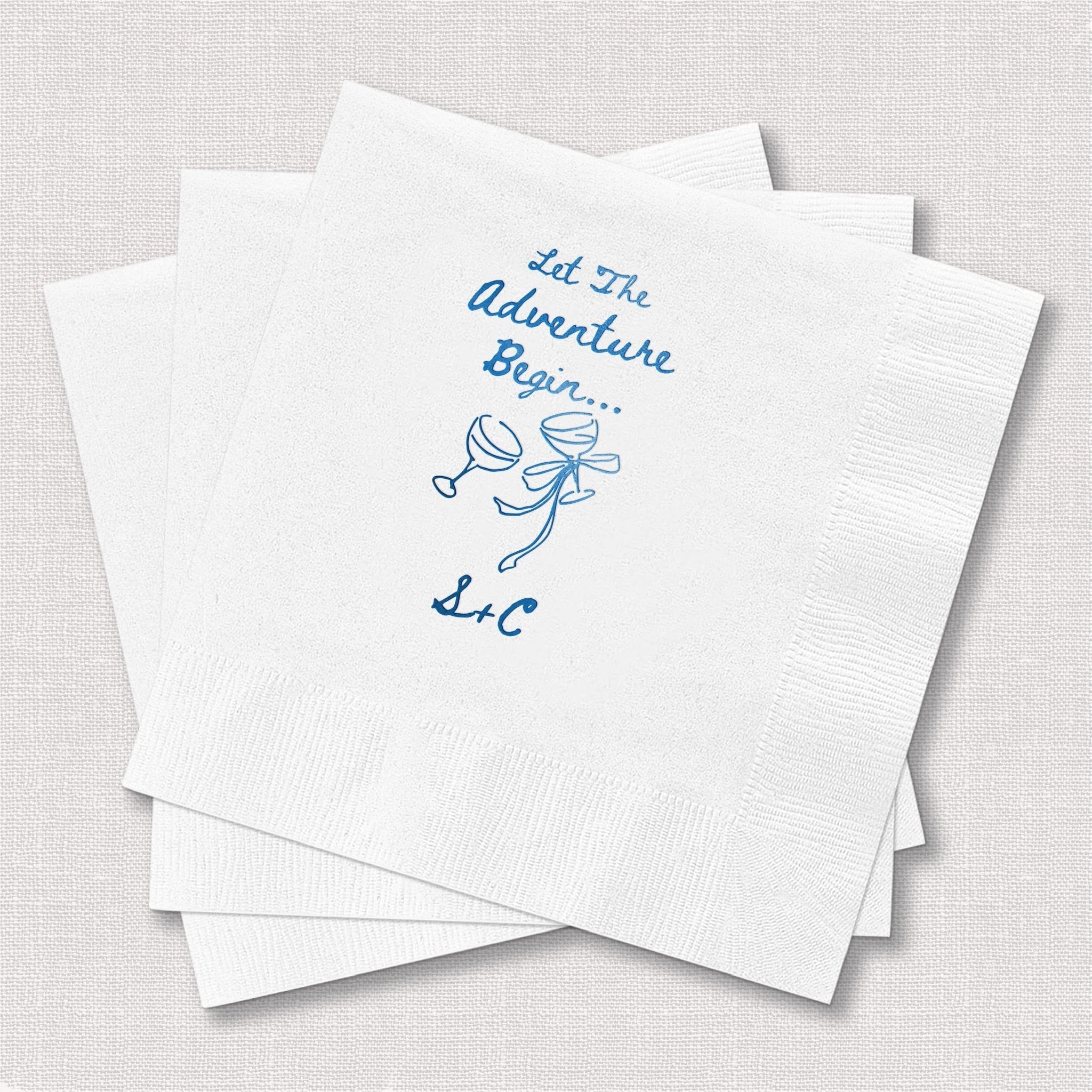 White cocktail napkins with blue foil-stamped cupid and "Let The Adventure Begin" text for weddings