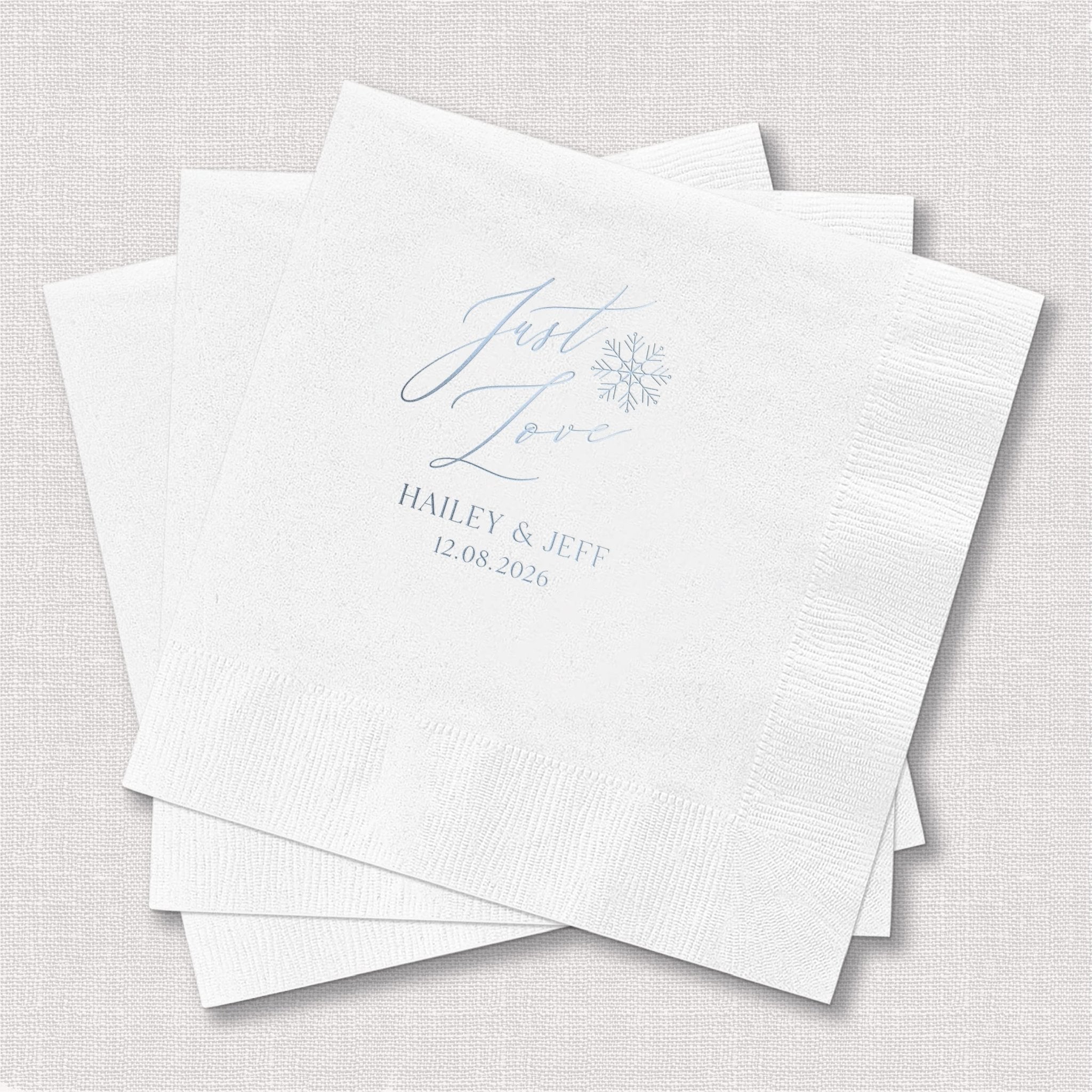 Stack of white napkins with blue foil-stamped "Just Love" text, snowflake design, and couple names Hailey and Jeff with wedding date 12.06.2020.