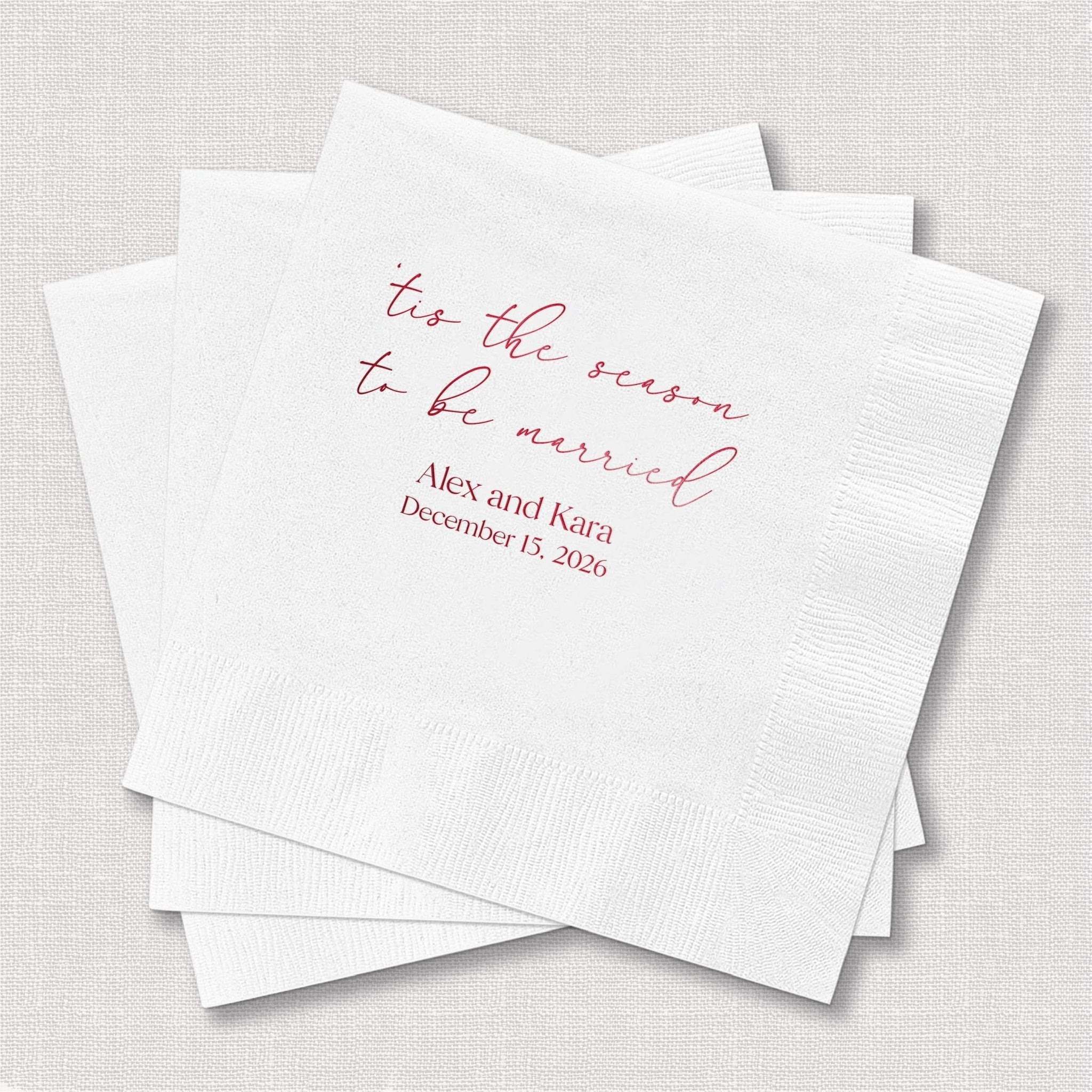 White cocktail napkins with pink foil-stamped text reading "tis the season to be married" and personalized names with wedding date