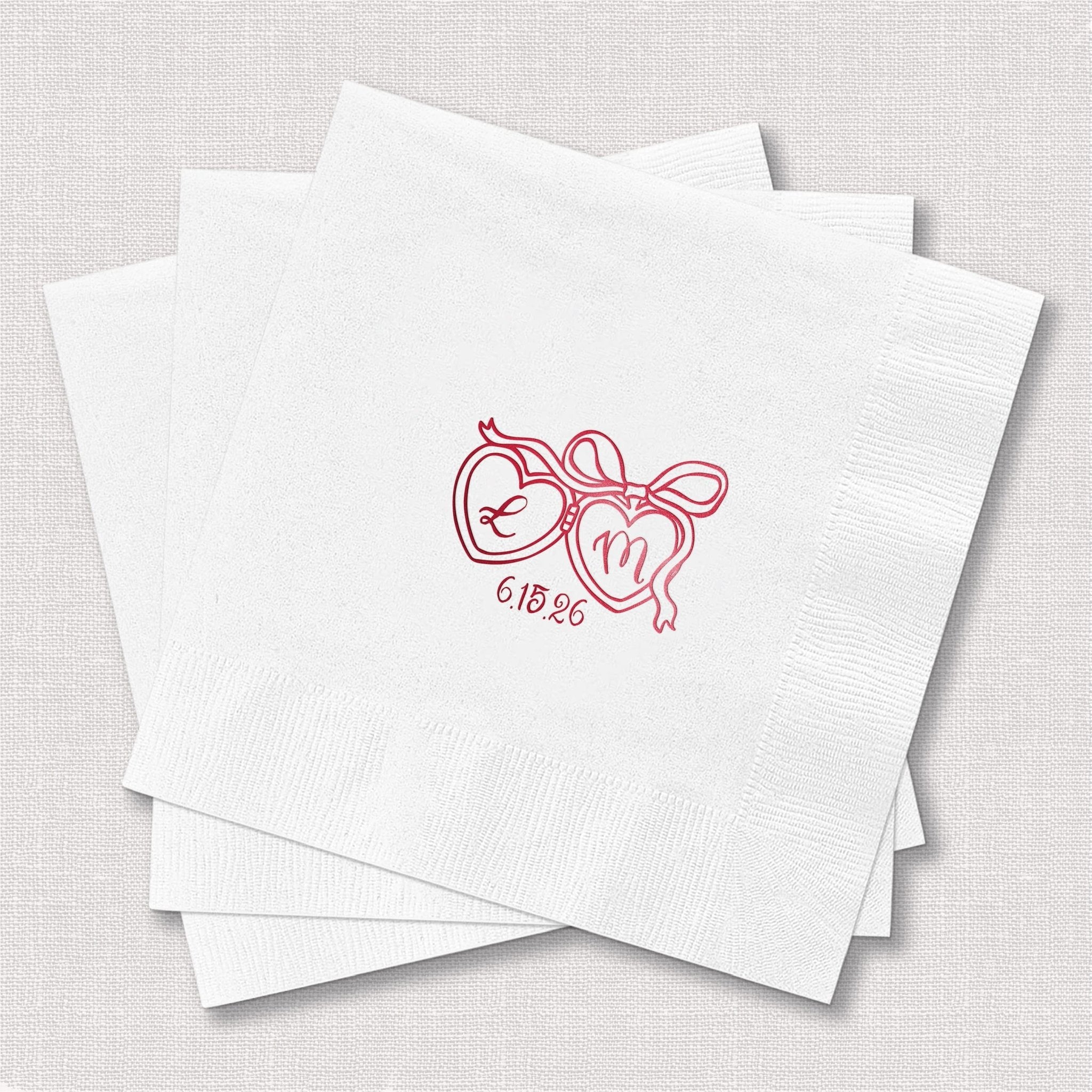 Stack of white cocktail napkins with pink foil-stamped heart locket design and initials "6.10.23"