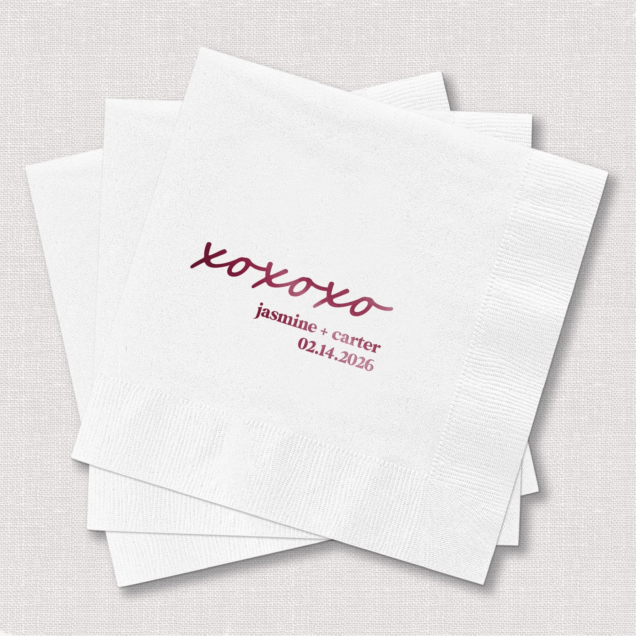 Stack of white cocktail napkins with burgundy foil-stamped xoxo design and personalized names and wedding date.