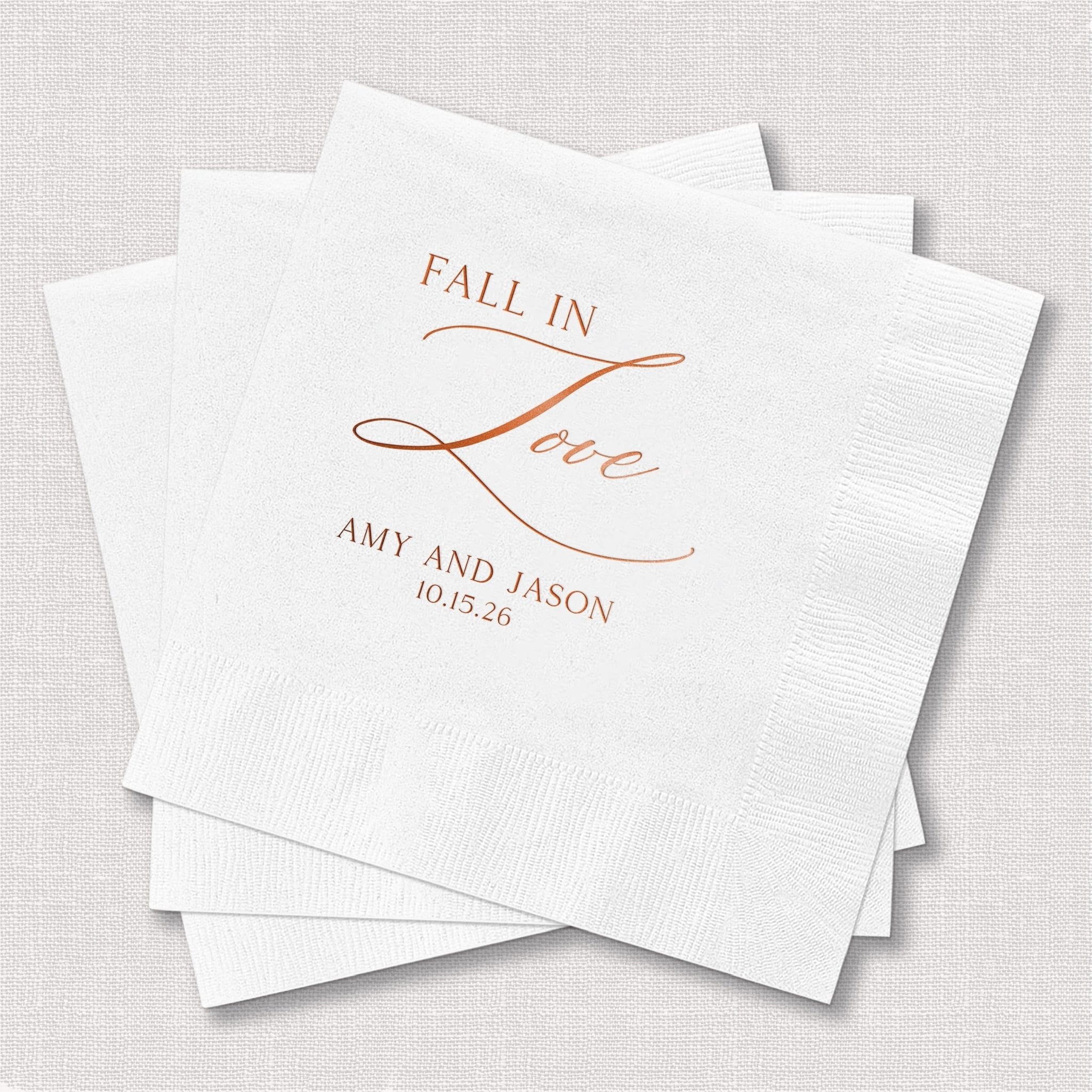 White napkins with copper foil-stamped text reading "Fall in Love" and personalized names and date for a wedding celebration.