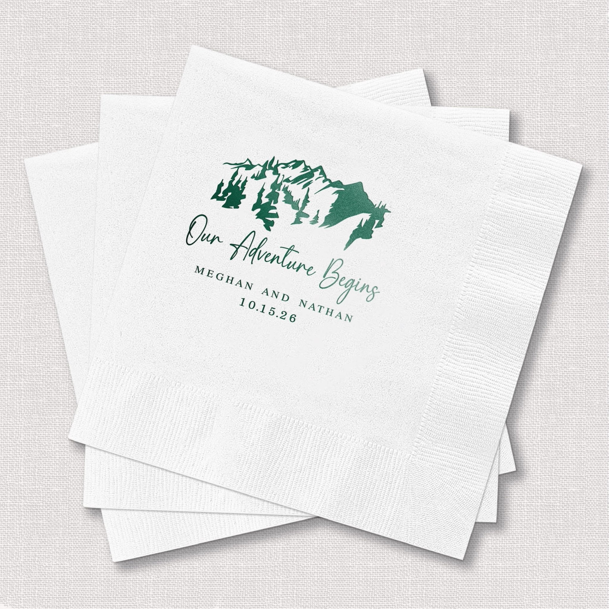 White cocktail napkins with green foil-stamped mountain map design and "Our Adventure Begins" text with personalized names and date.