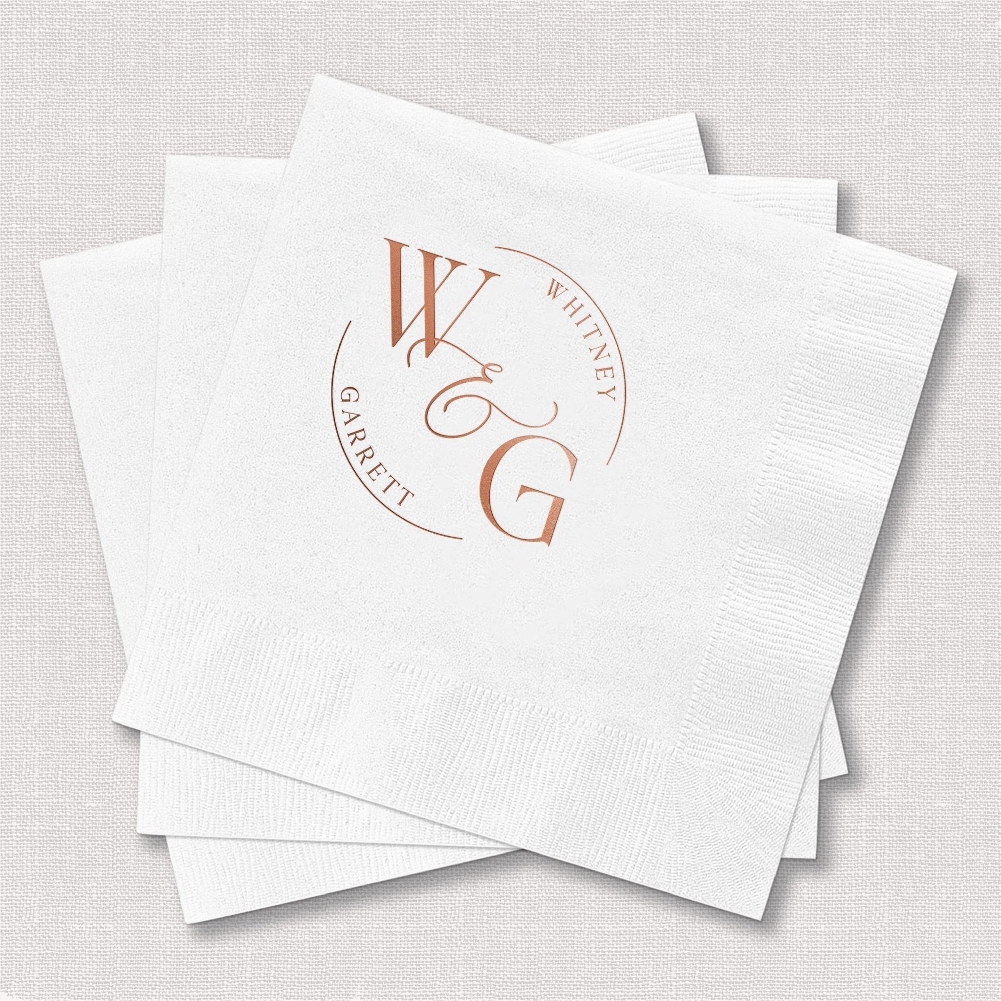 White cocktail napkins with rose gold foil-stamped circular monogram design featuring initials and wedding date.