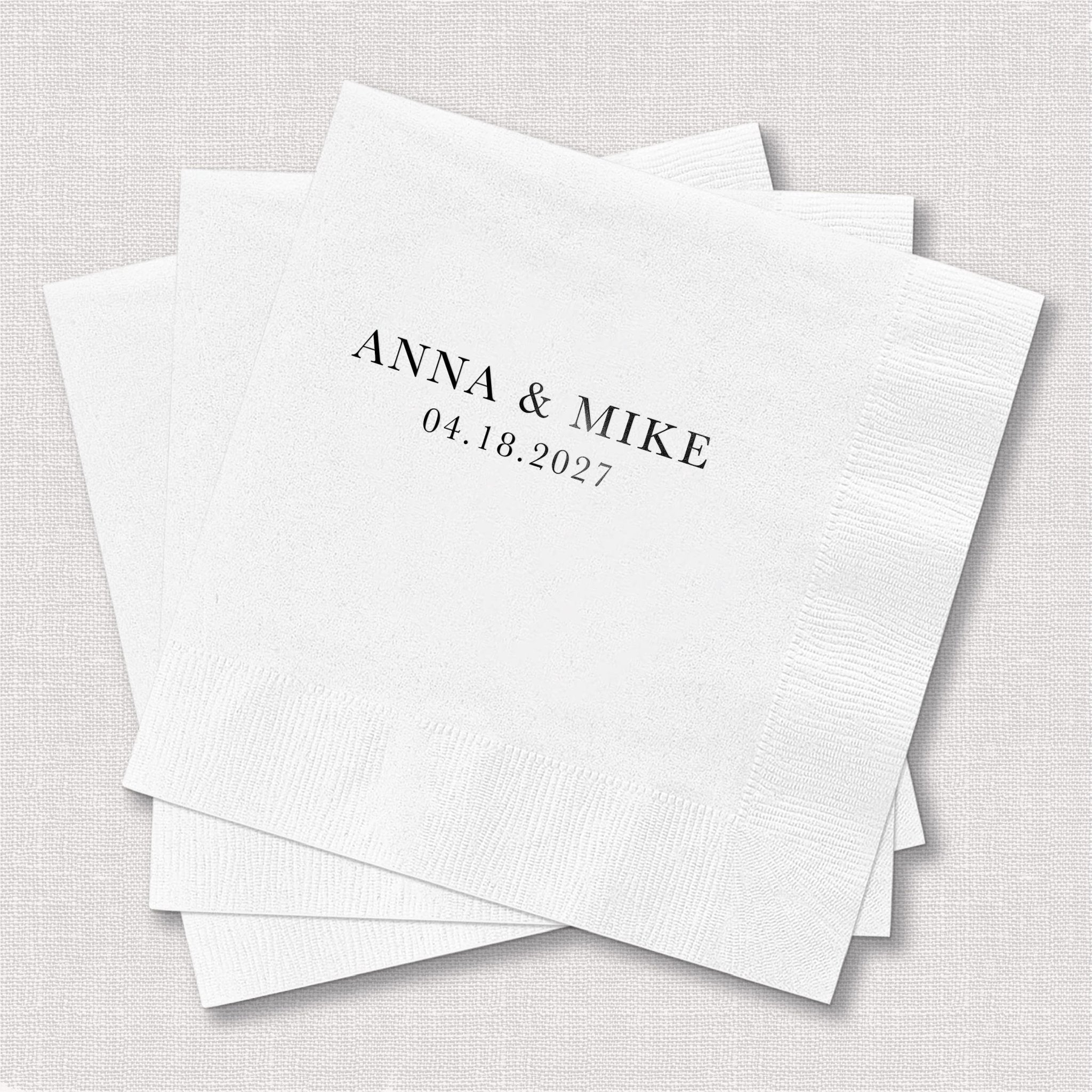 White cocktail napkins with "ANNA & MIKE 04.18.2027" in foil stamped text, stacked in a pile
