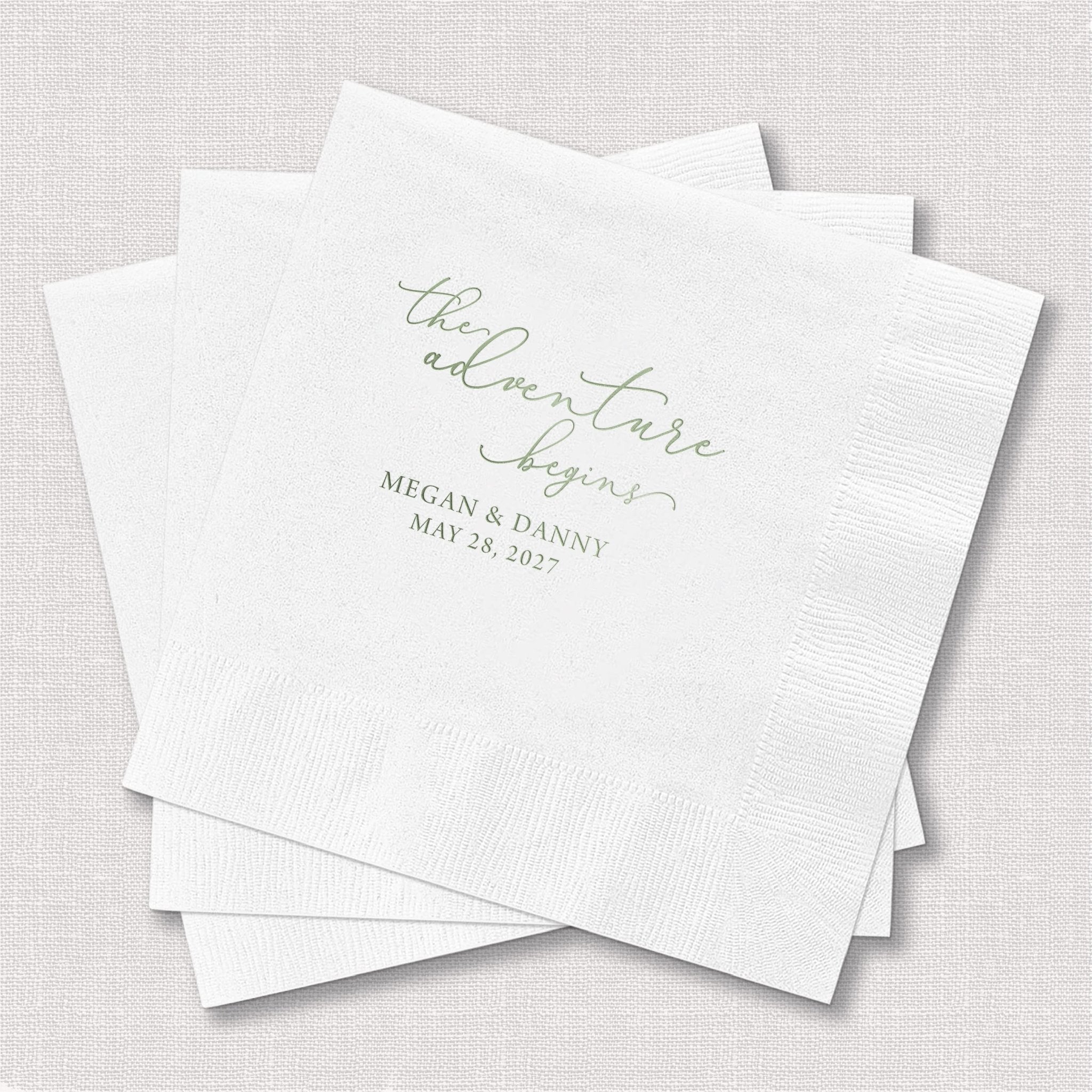 Stack of white napkins with "the adventure begins" in script and couple names and date in foil stamped text