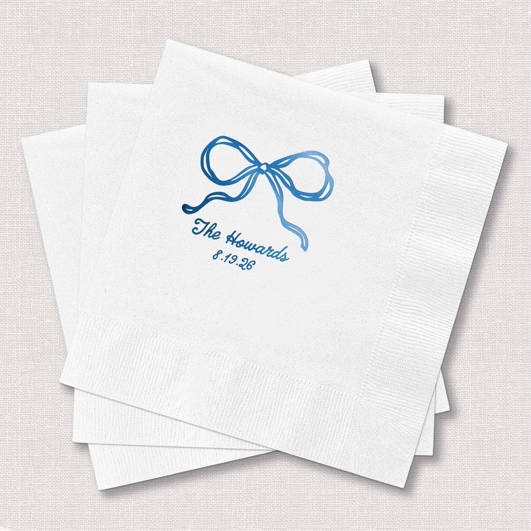 Stack of white cocktail napkins with blue foil-stamped bow and personalized text reading "The Hammonds 7.19.24"