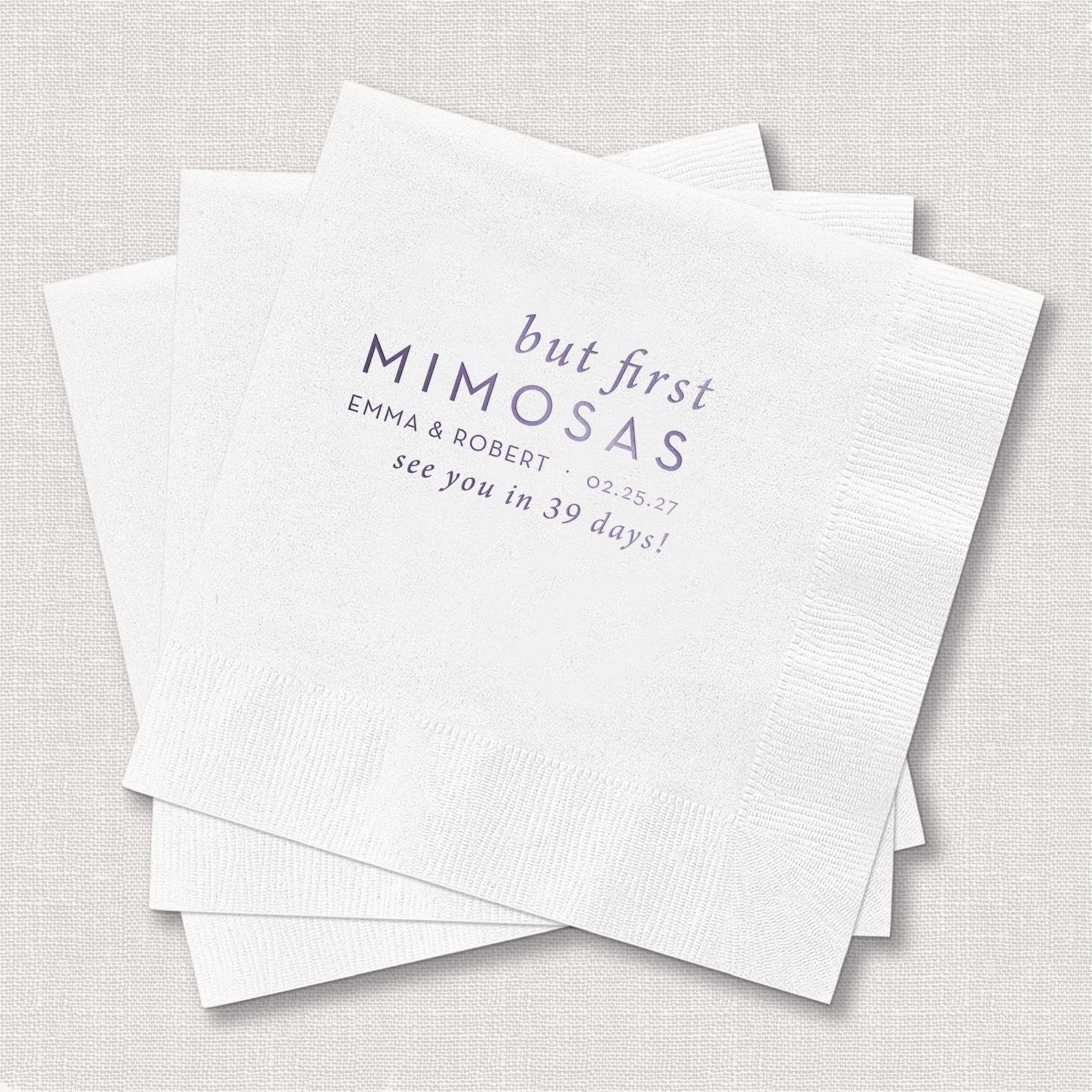 White cocktail napkins with "but first MIMOSAS" text and personalized names in foil stamping
