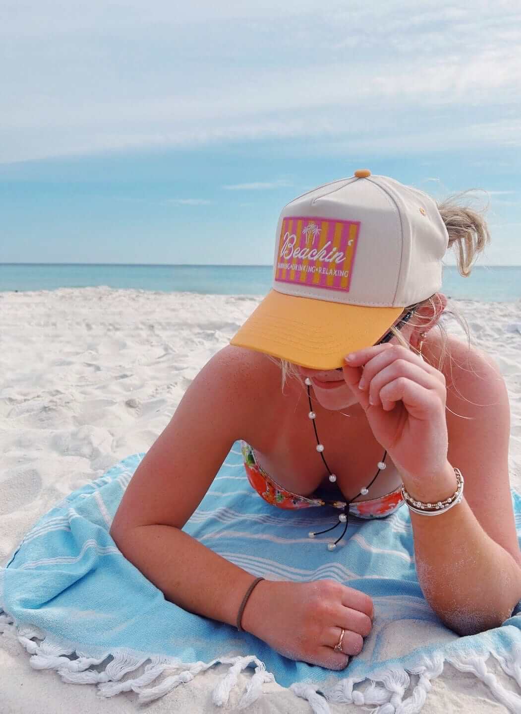 White and yellow trucker hat with pink "Beachin'" patch worn on a beach with ocean waves in background.
