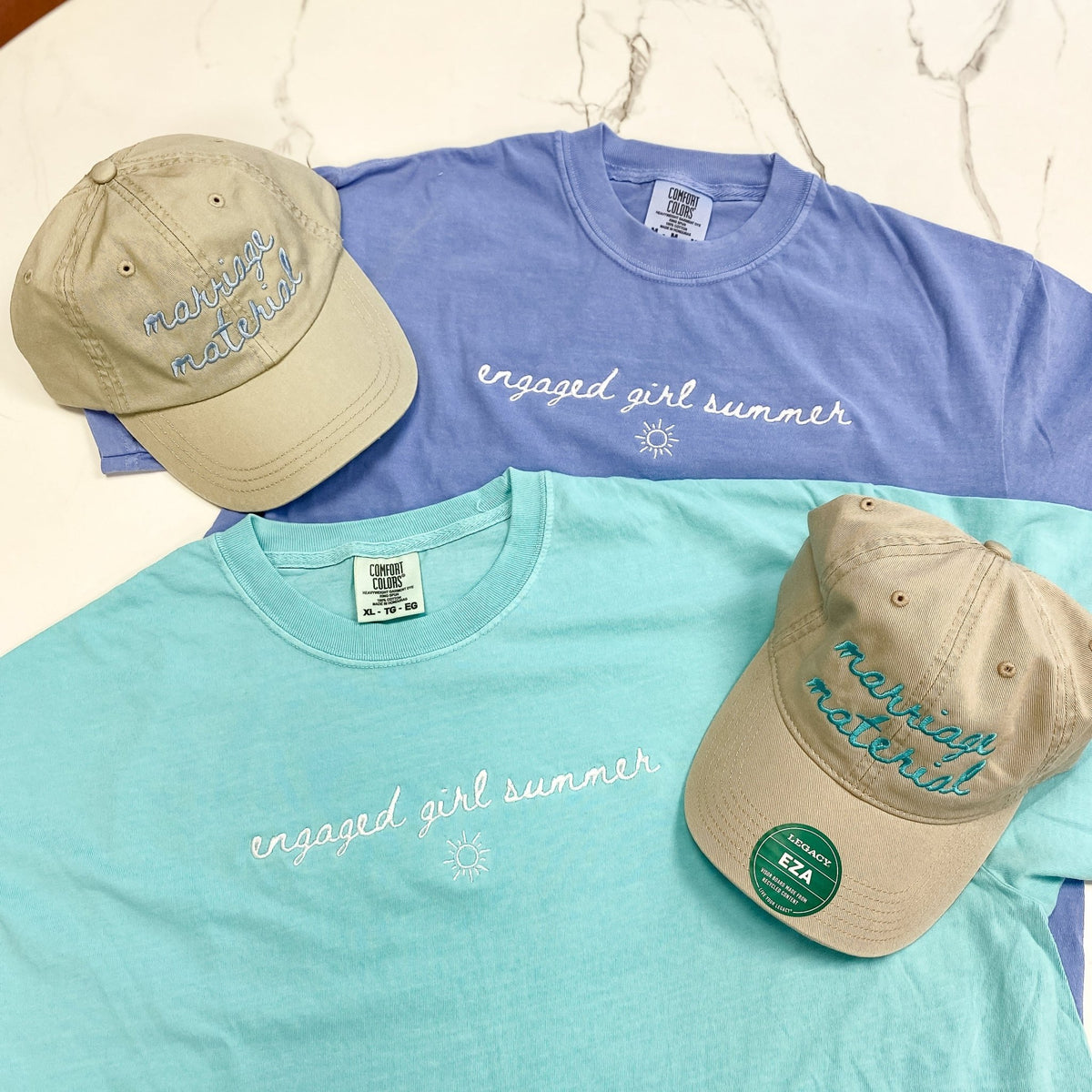 Blue and mint green t-shirts with "engaged girl summer" script text, displayed with matching embroidered baseball caps in cream and tan.