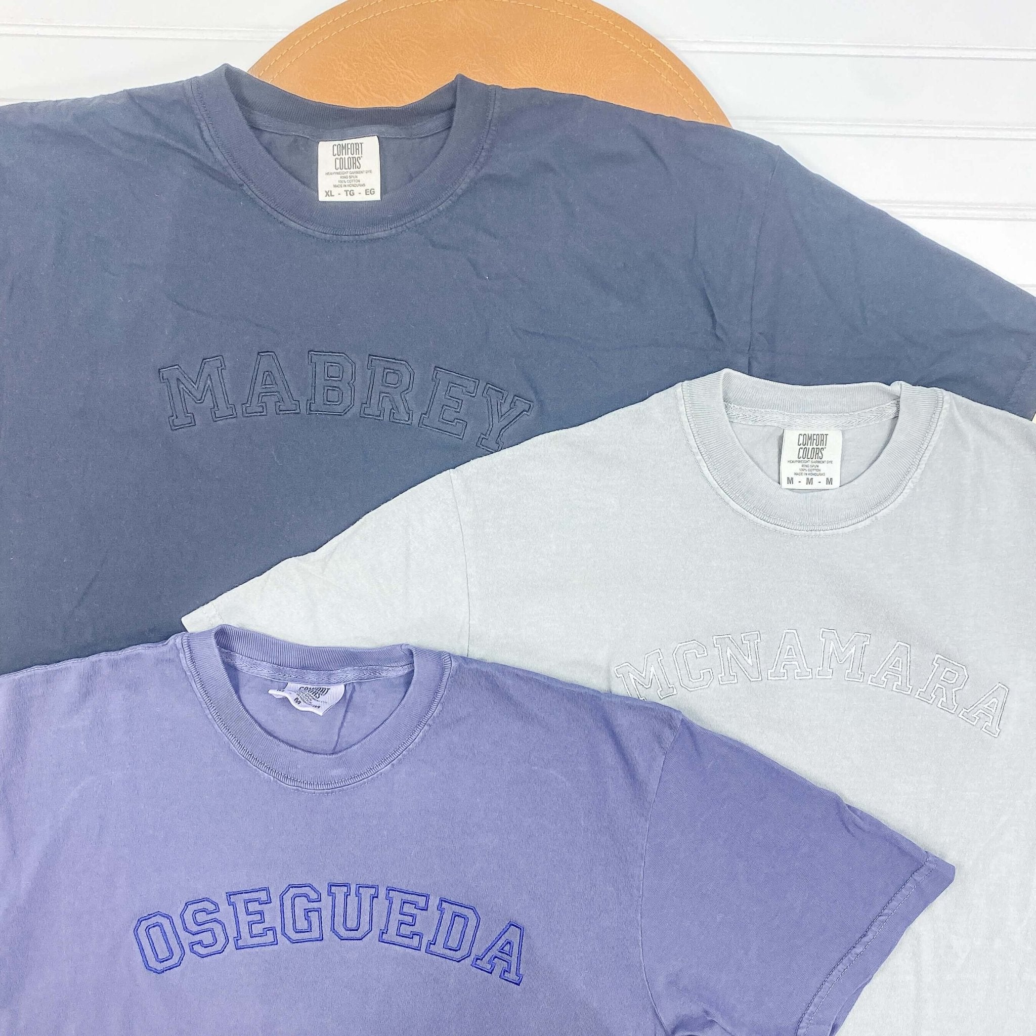 Three stacked personalized t-shirts in slate blue, cream, and lavender with embroidered last names displayed on wooden hanger.