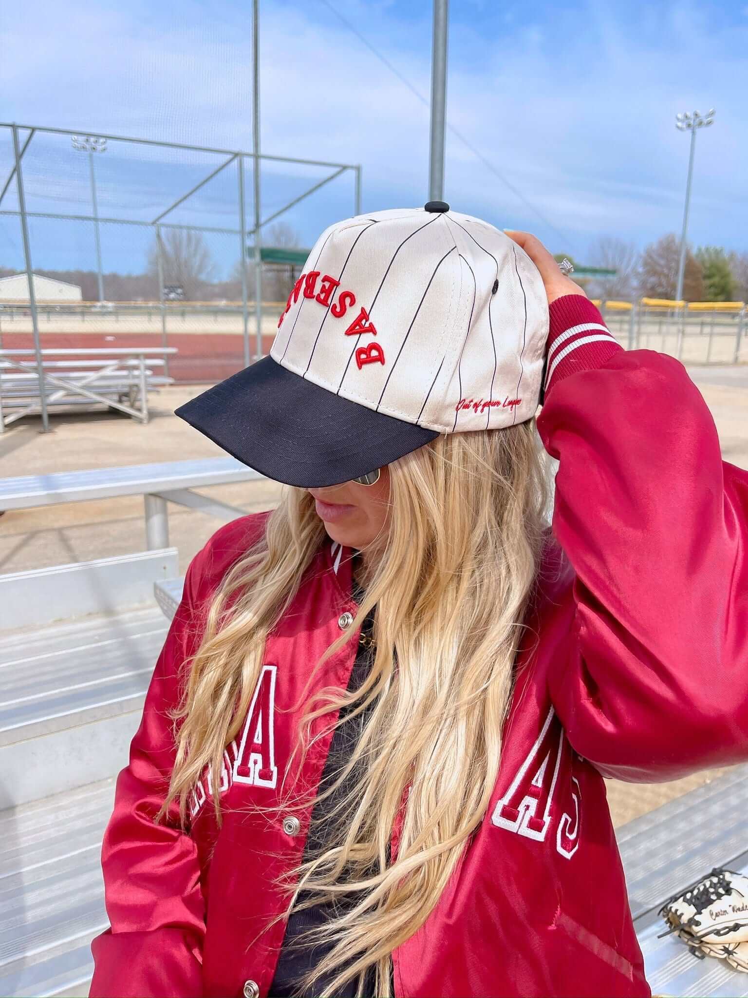 White and navy baseball cap with red vintage text and pinstripe design, worn with red jacket at baseball field