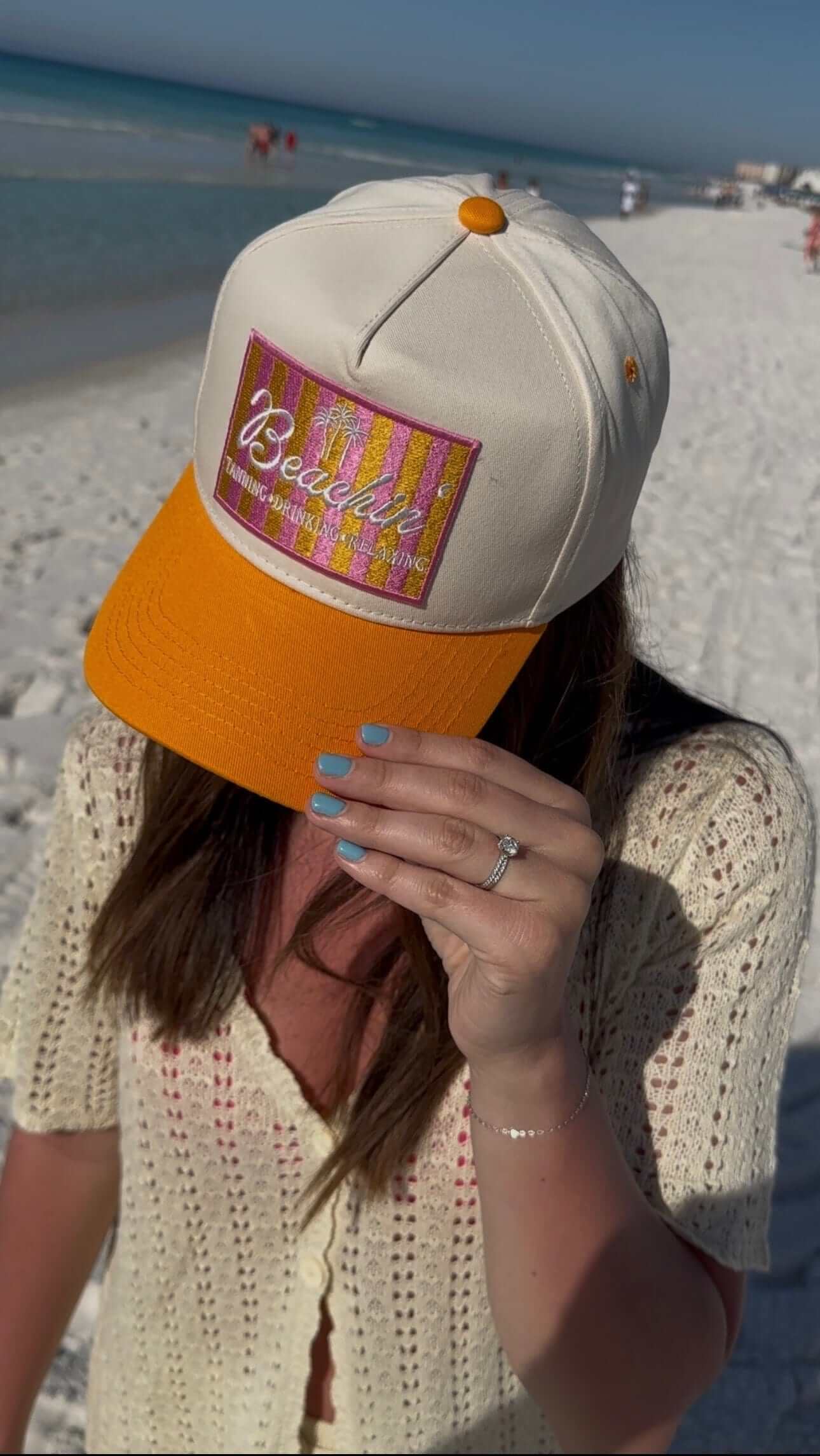White and orange trucker hat with embroidered "Beachin'" patch, worn at the beach with ocean in background.