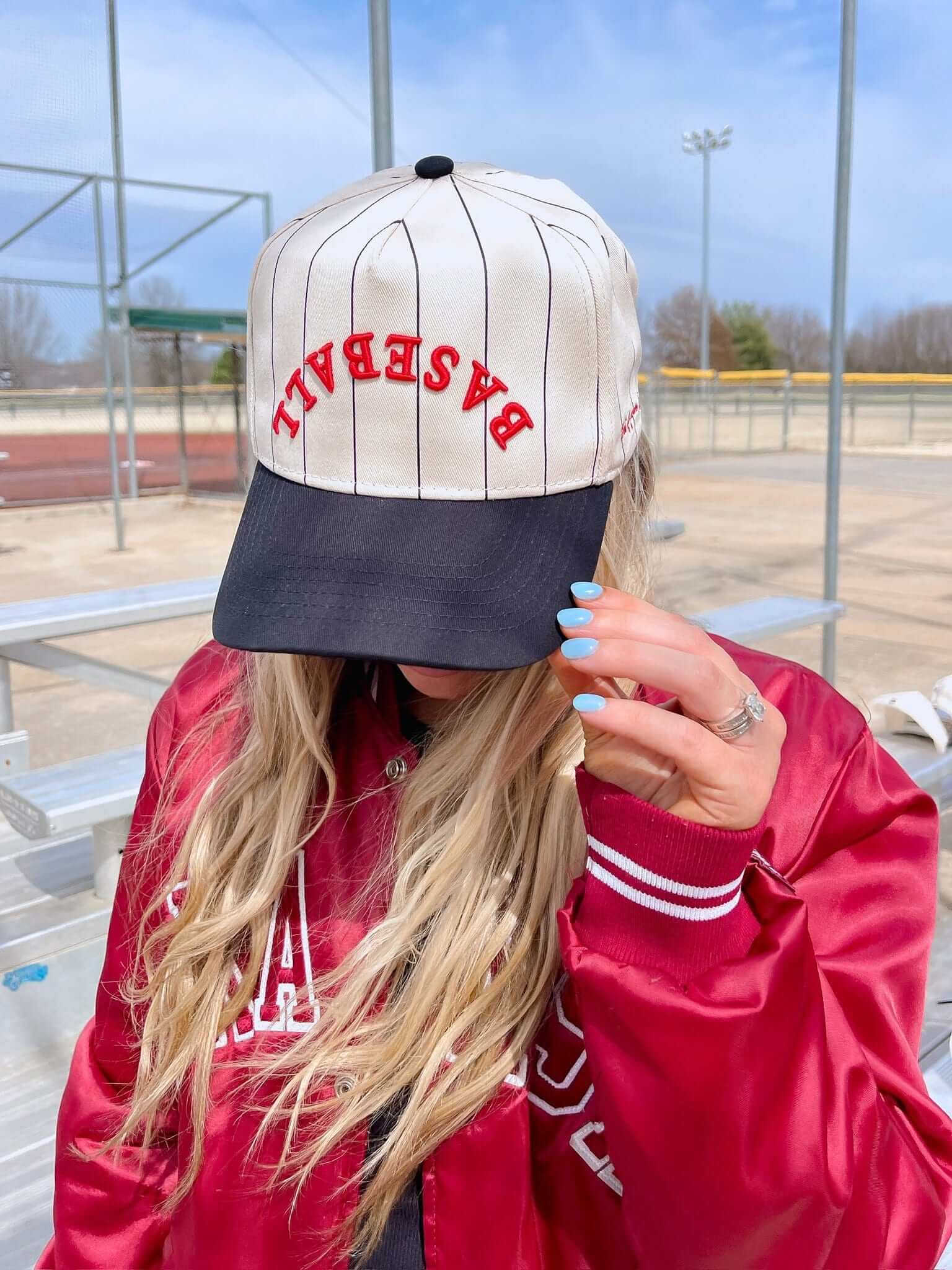 Cream pinstriped baseball cap with red "DYNASTY" text and navy blue bill, worn by person in pink jacket at baseball field.