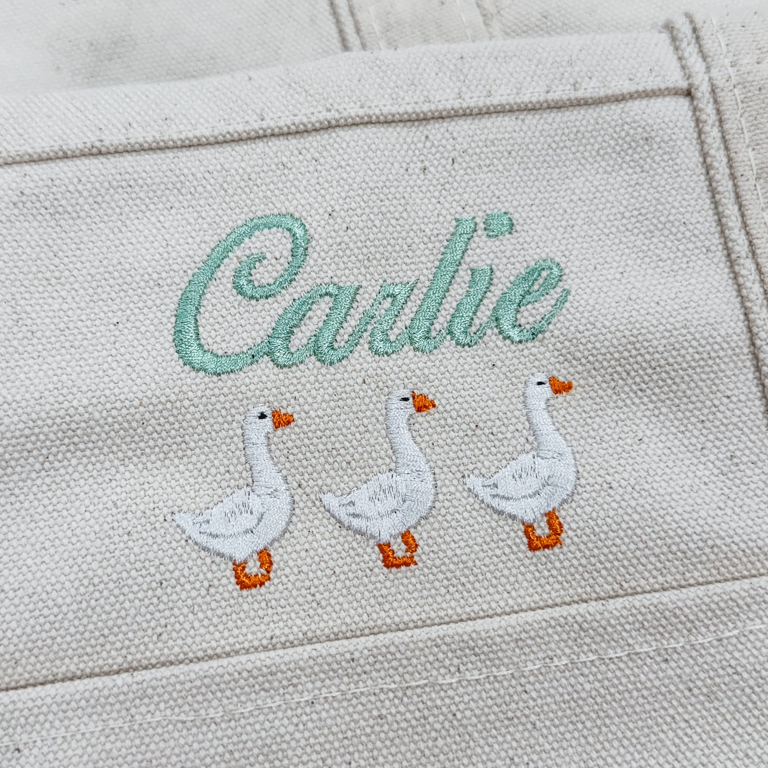 Close-up of custom name embroidery and three white geese on a natural canvas tote bag