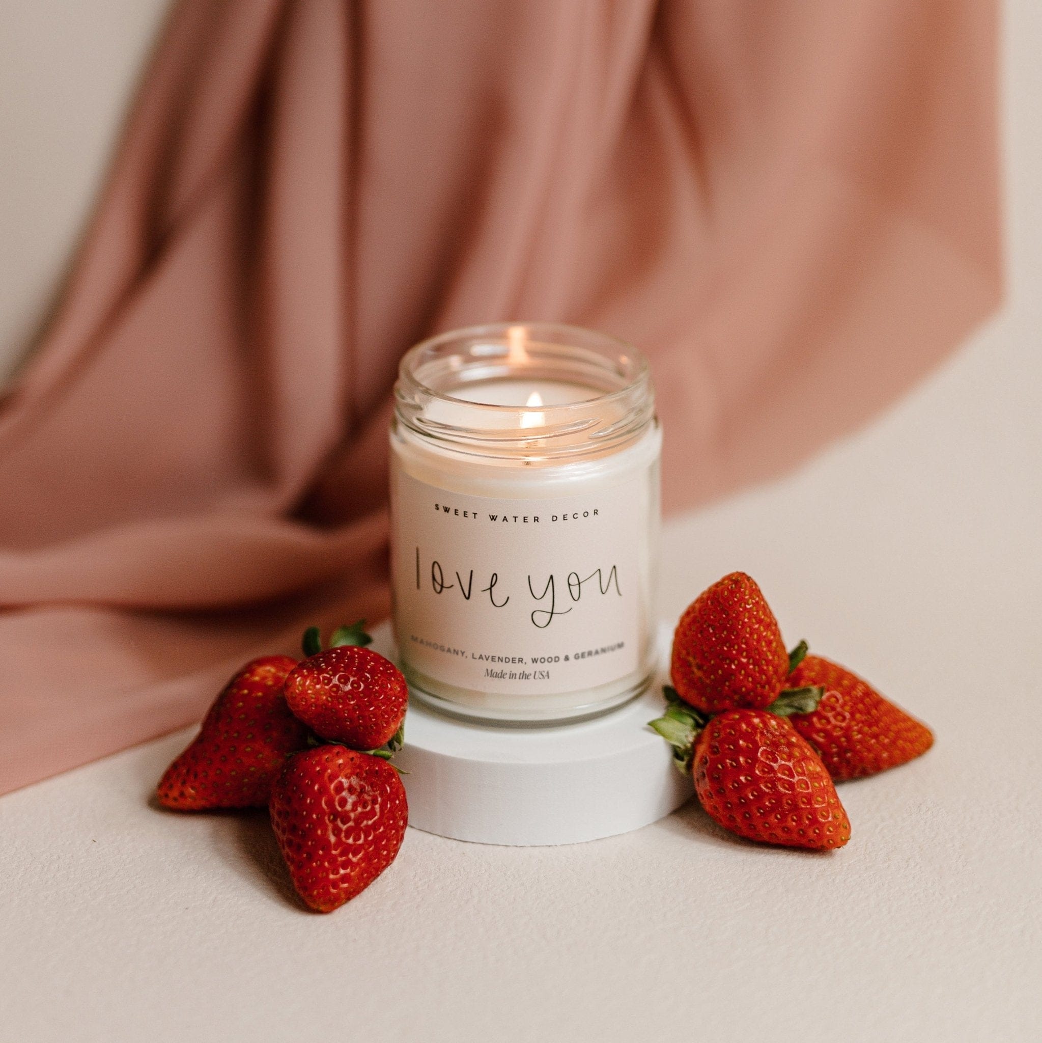 Clear soy candle jar with "love you" text, surrounded by fresh strawberries and draped blush fabric.