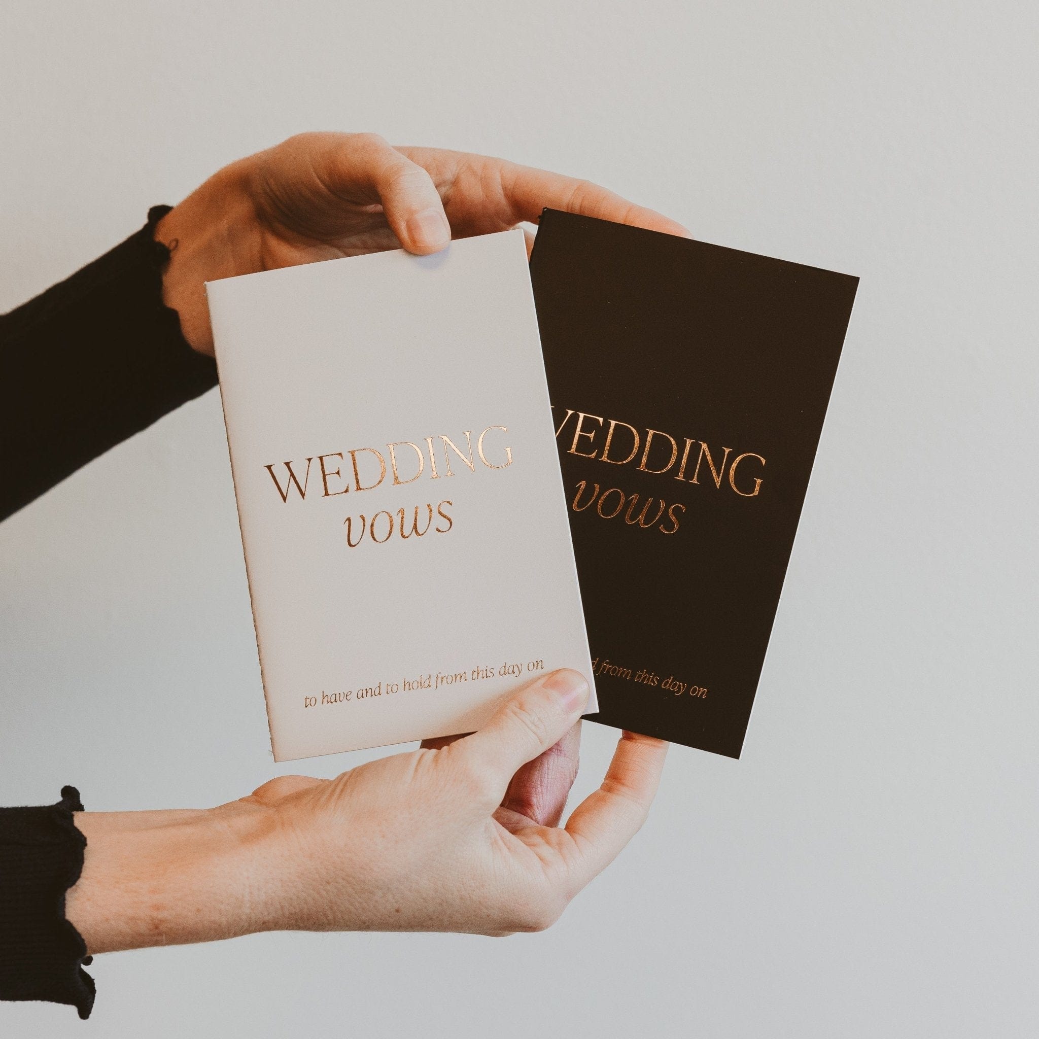 Hand holding open white and black wedding vows journals with gold foil text on cream background