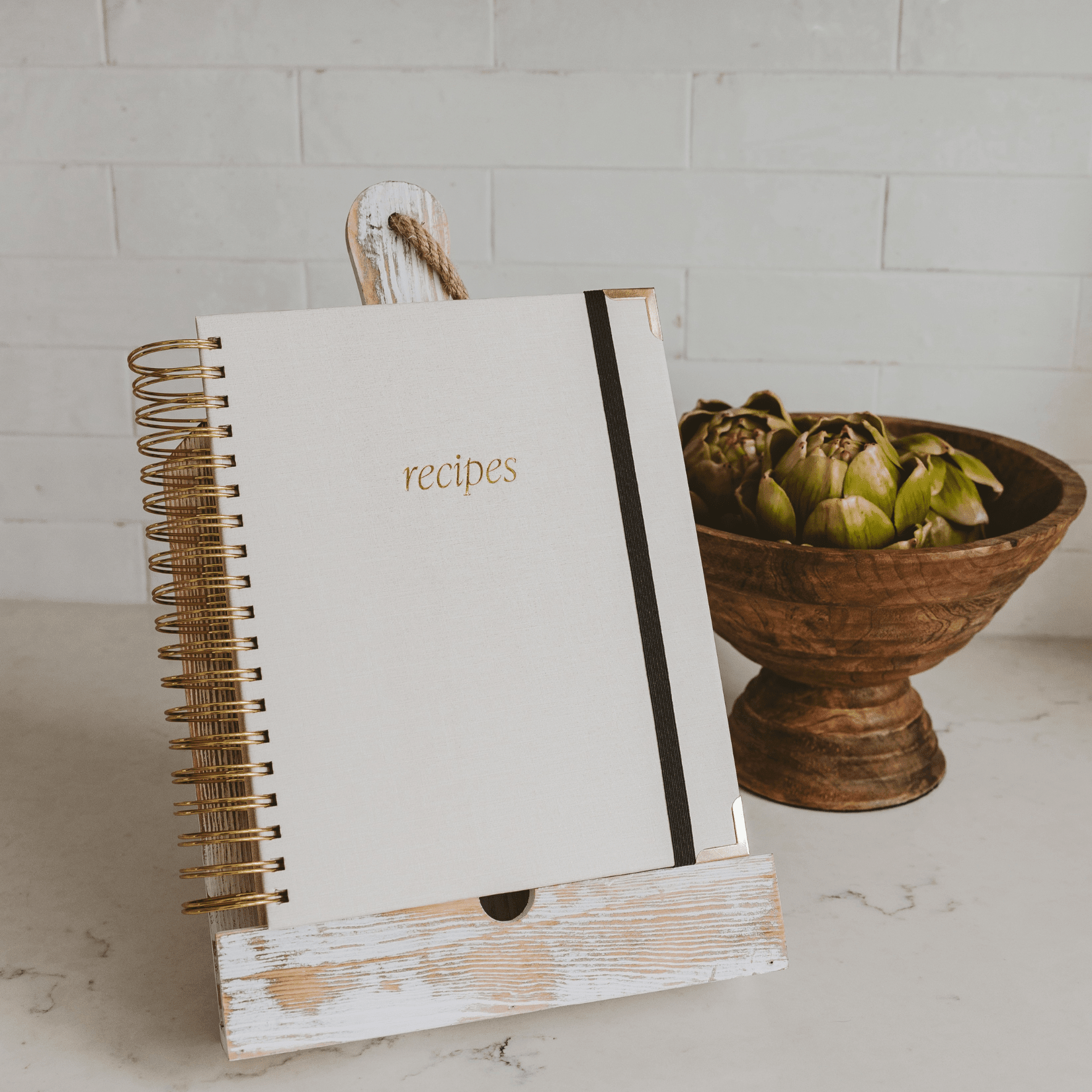 White spiral-bound recipe book with gold "recipes" text and black elastic band closure, displayed in whitewashed wooden stand.