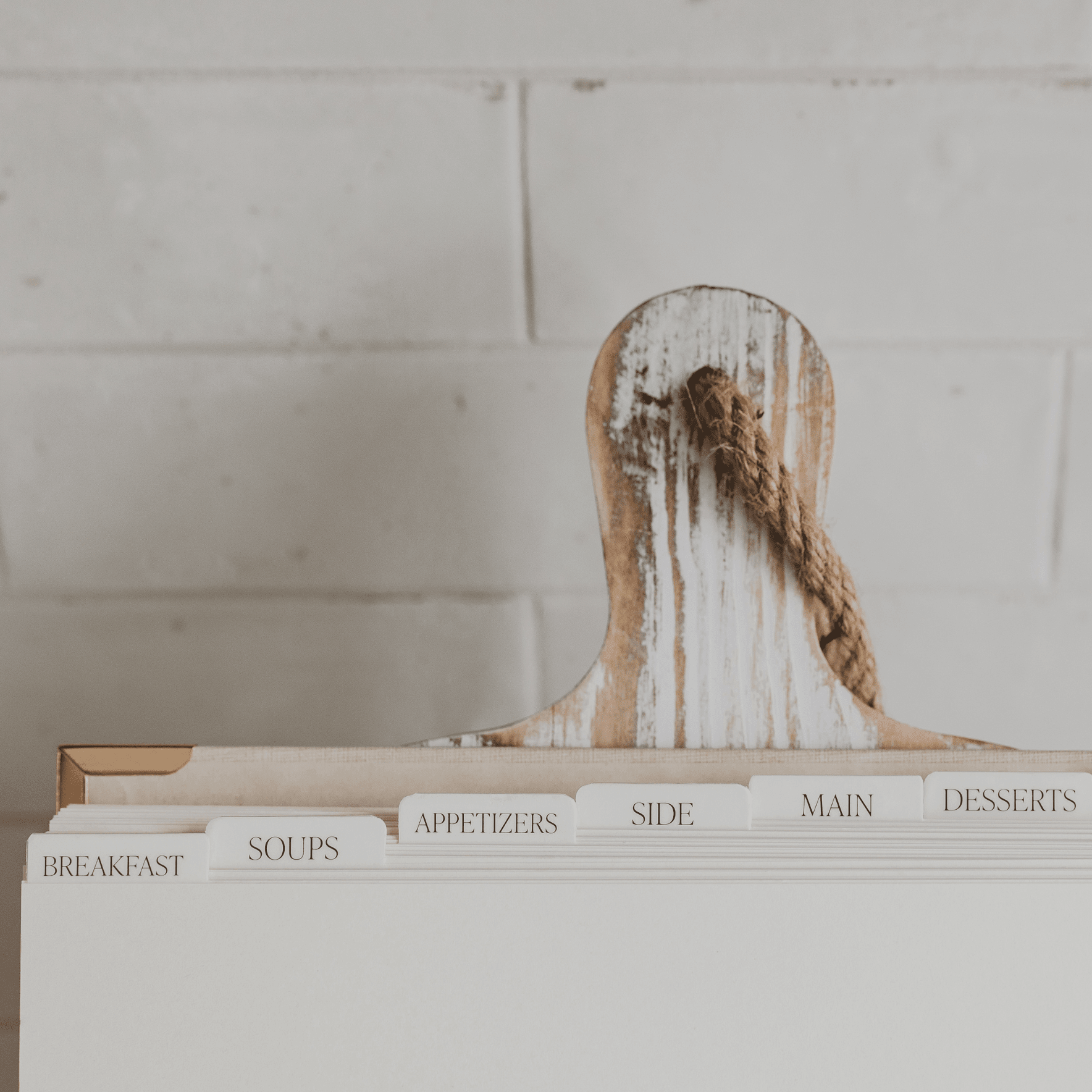 Whitewashed wooden recipe book organizer with meal category dividers including breakfast, soups, appetizers, side, main, and desserts, featuring a rope handle.