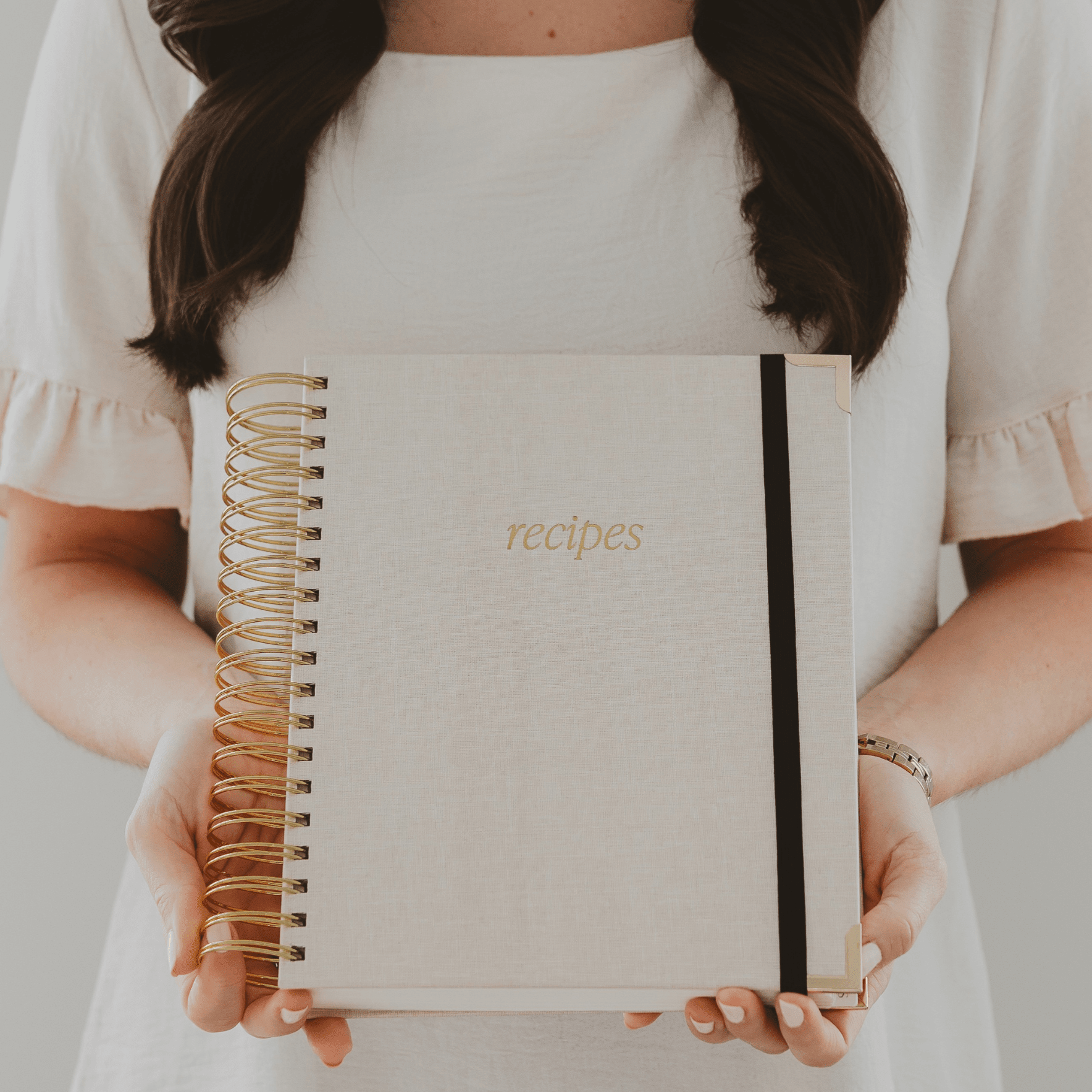 Cream spiral-bound recipe book with gold foil "recipes" text and black elastic closure band, held by person in white shirt.