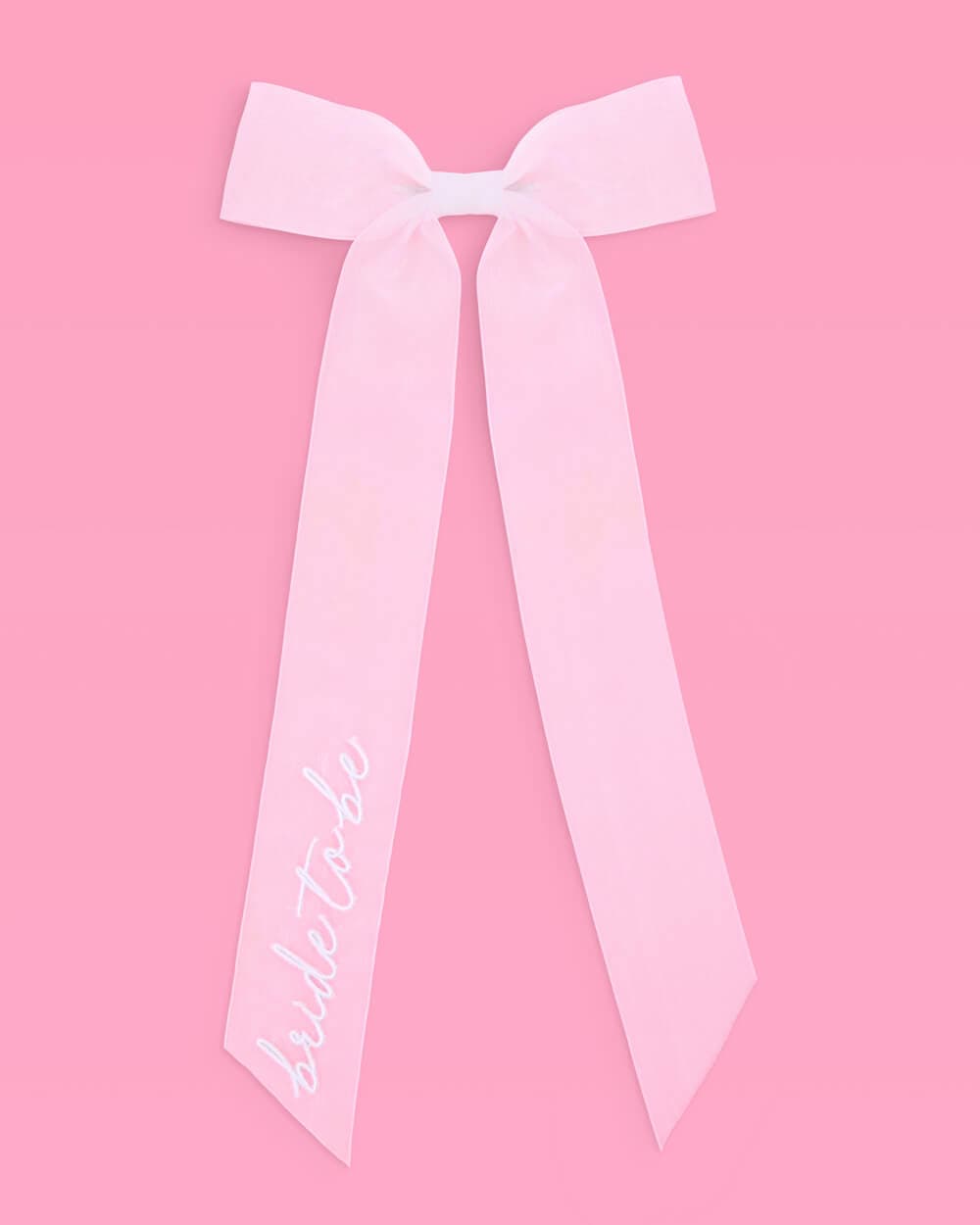 White satin bow with "bride to be" embroidered script on left tail, displayed against pink background.