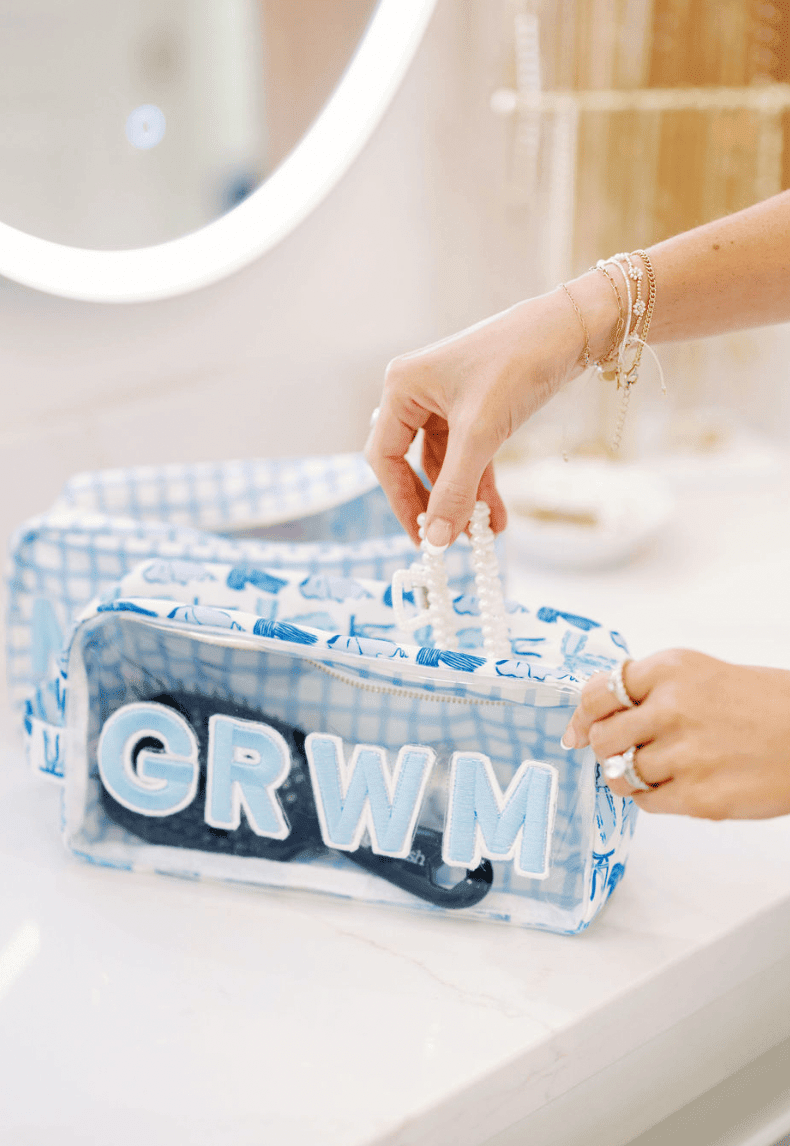 Clear makeup bag with blue gingham pattern and "GRWM" text, held by hands with bracelets