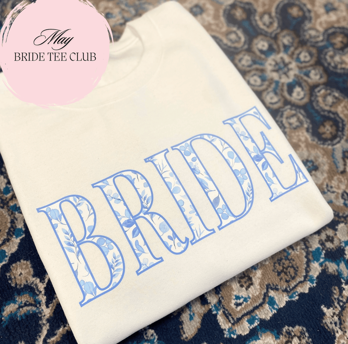 Cream colored t-shirt with blue "BRIDE" text in decorative lettering, folded on patterned rug background