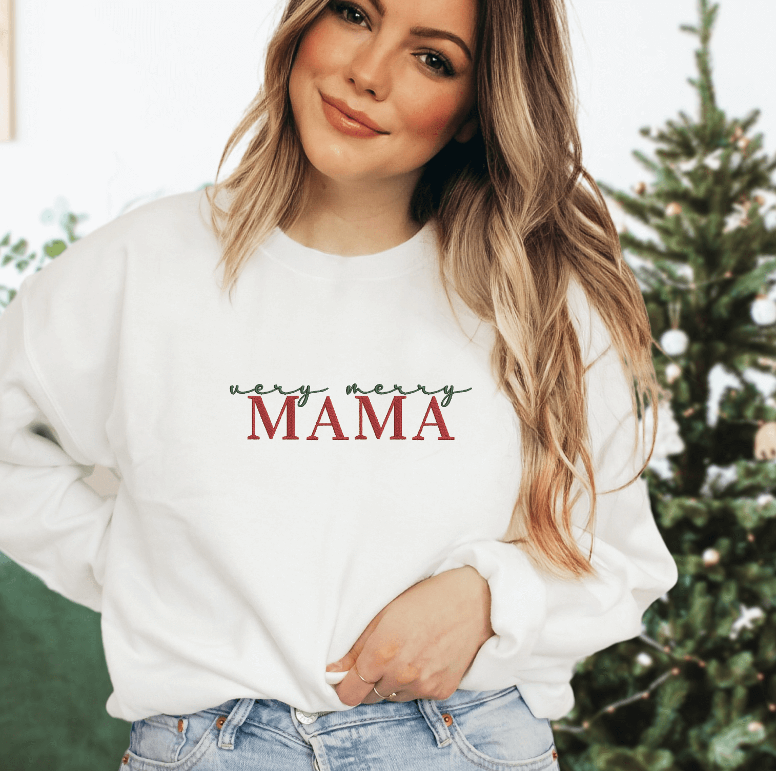 Cream sweatshirt with red embroidered "MAMA" text featuring holly leaf design, worn by woman in front of decorated Christmas tree.