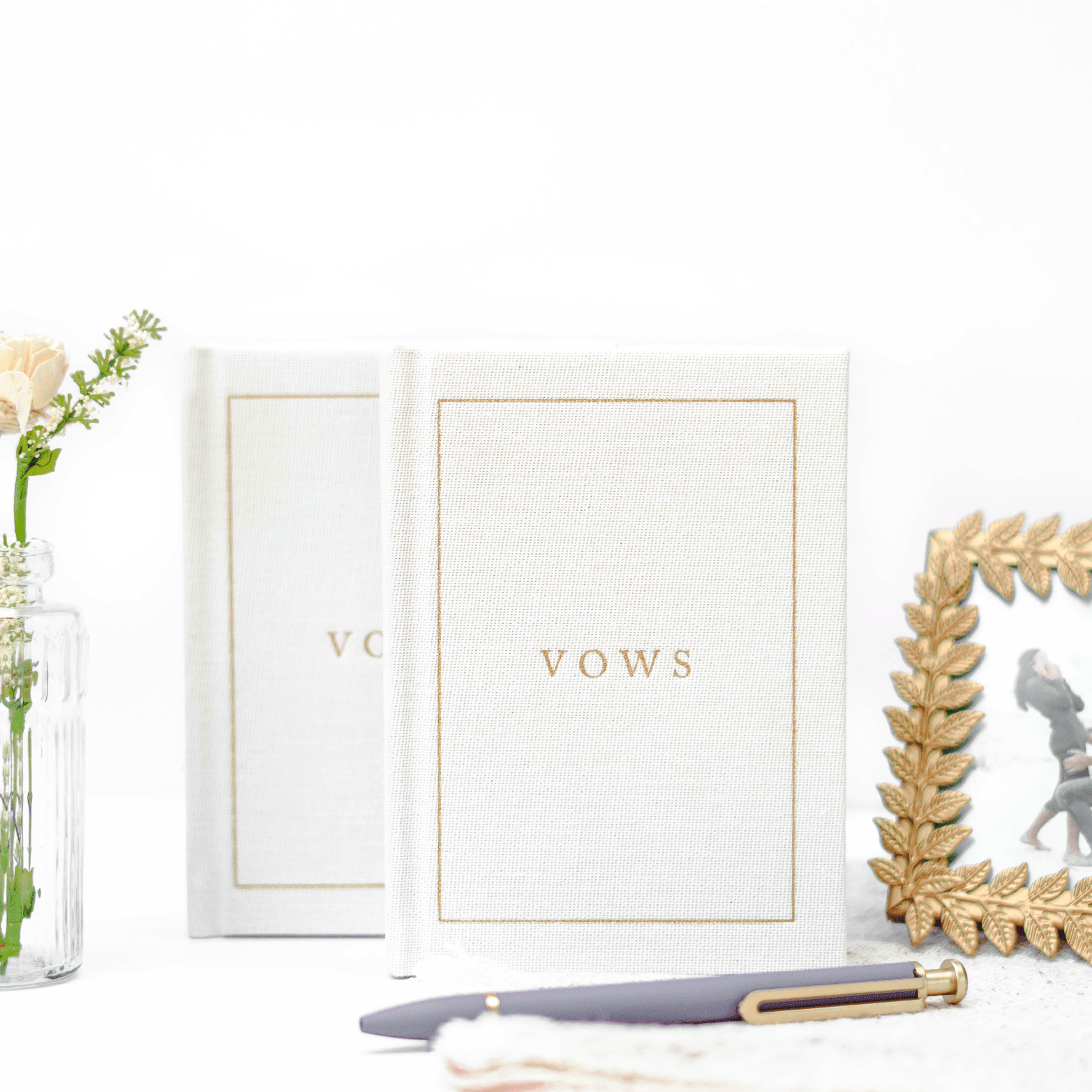 Cream vow books with gold text and border, displayed with flowers and gold frame accents