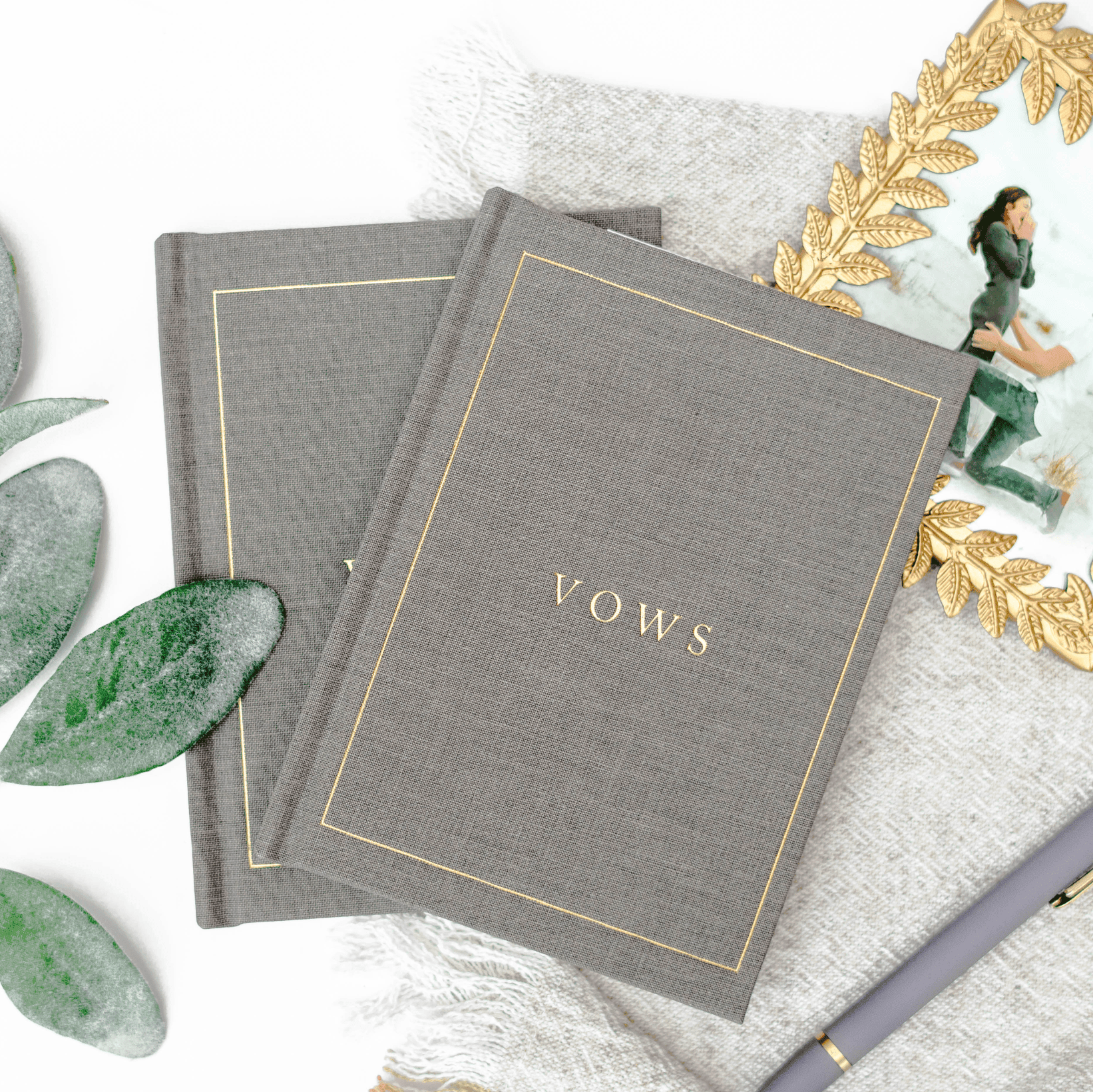 Gray linen vow books with gold foil "VOWS" text, displayed on white fabric with eucalyptus leaves and gold accents.