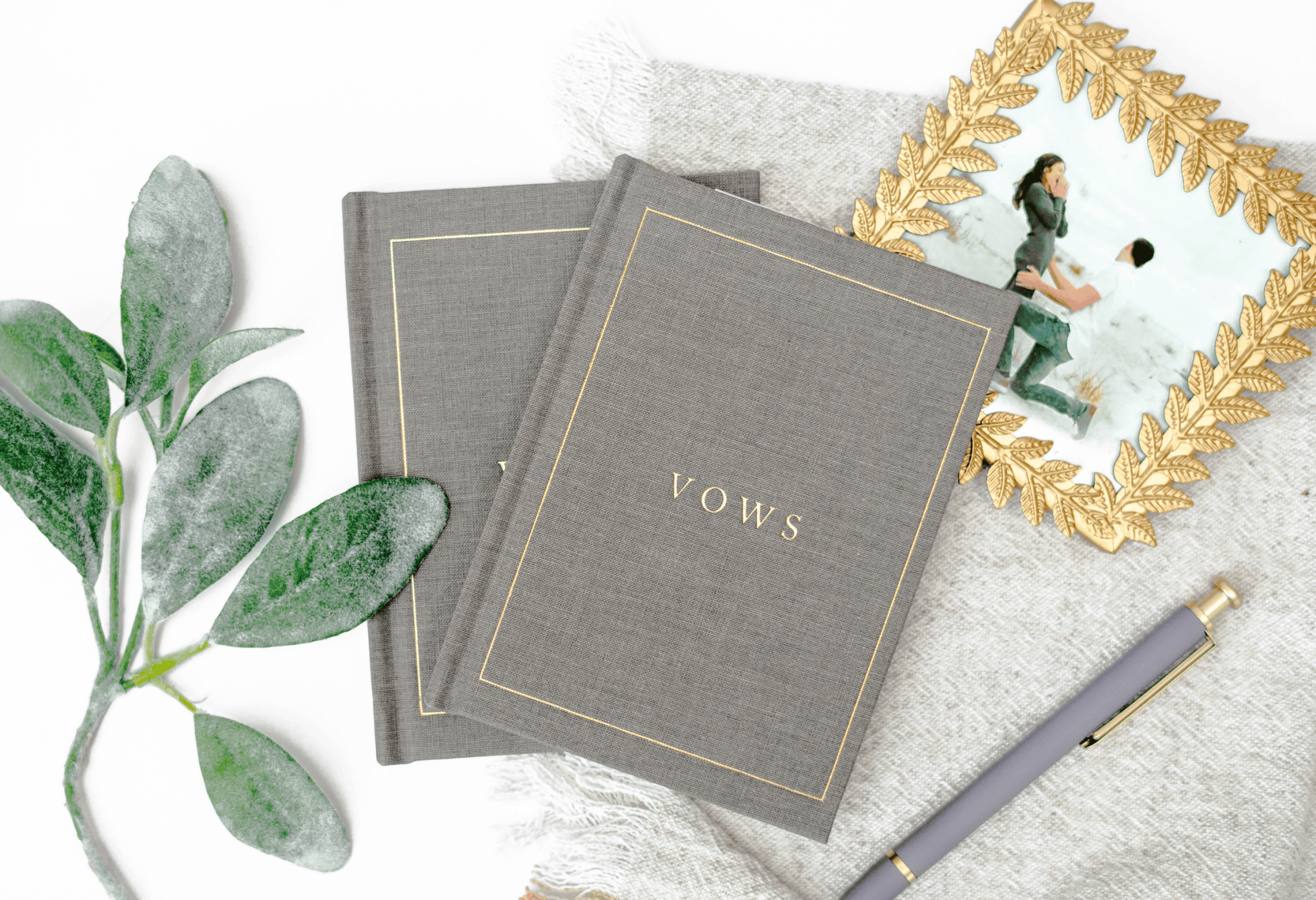 Gray linen vow books with gold "VOWS" text, styled with greenery, a framed photo, and pen on white fabric.