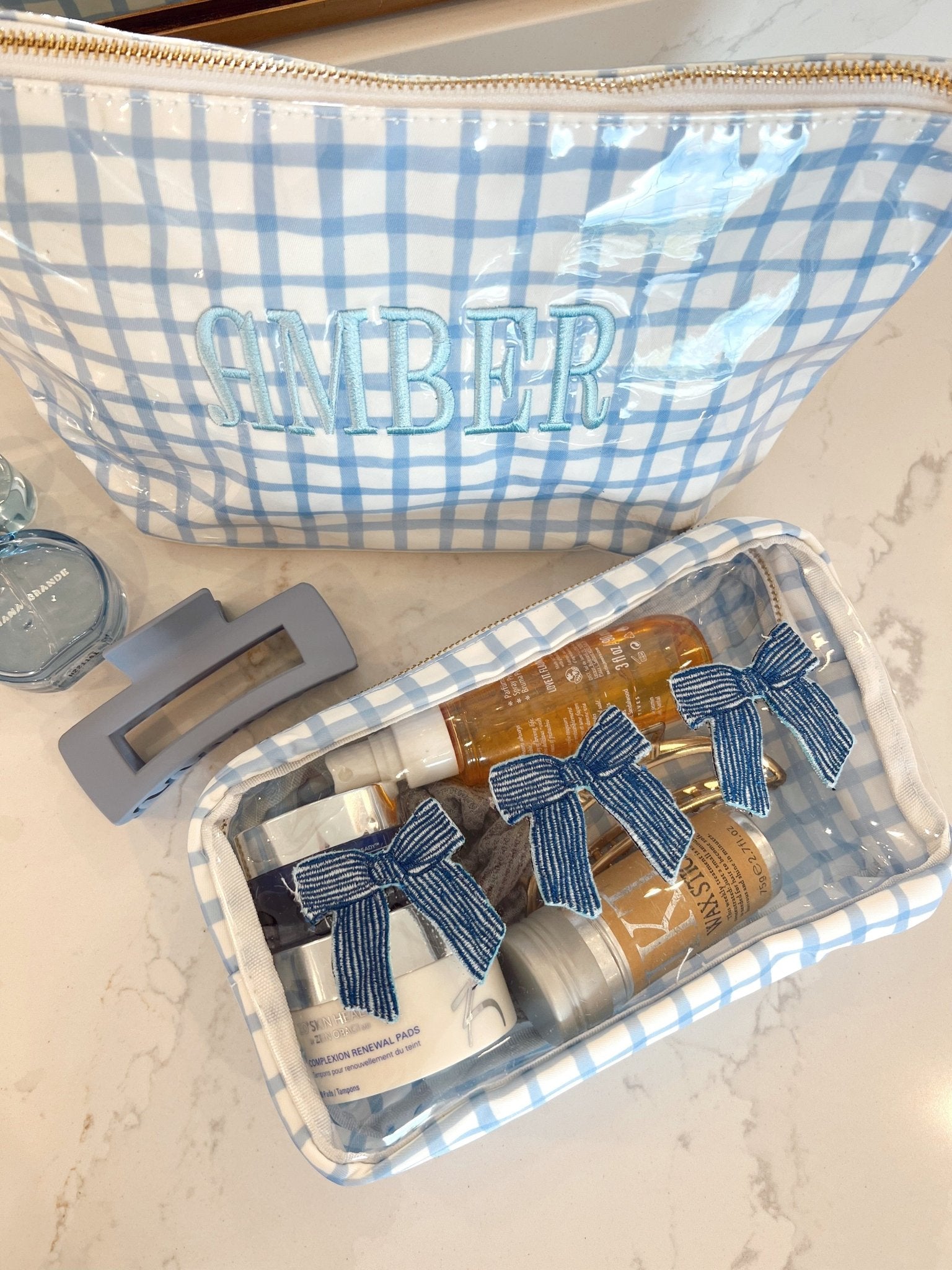 Blue gingham makeup bag personalized with "AMBER" text and matching clear case with blue striped bows containing cosmetics and brushes.