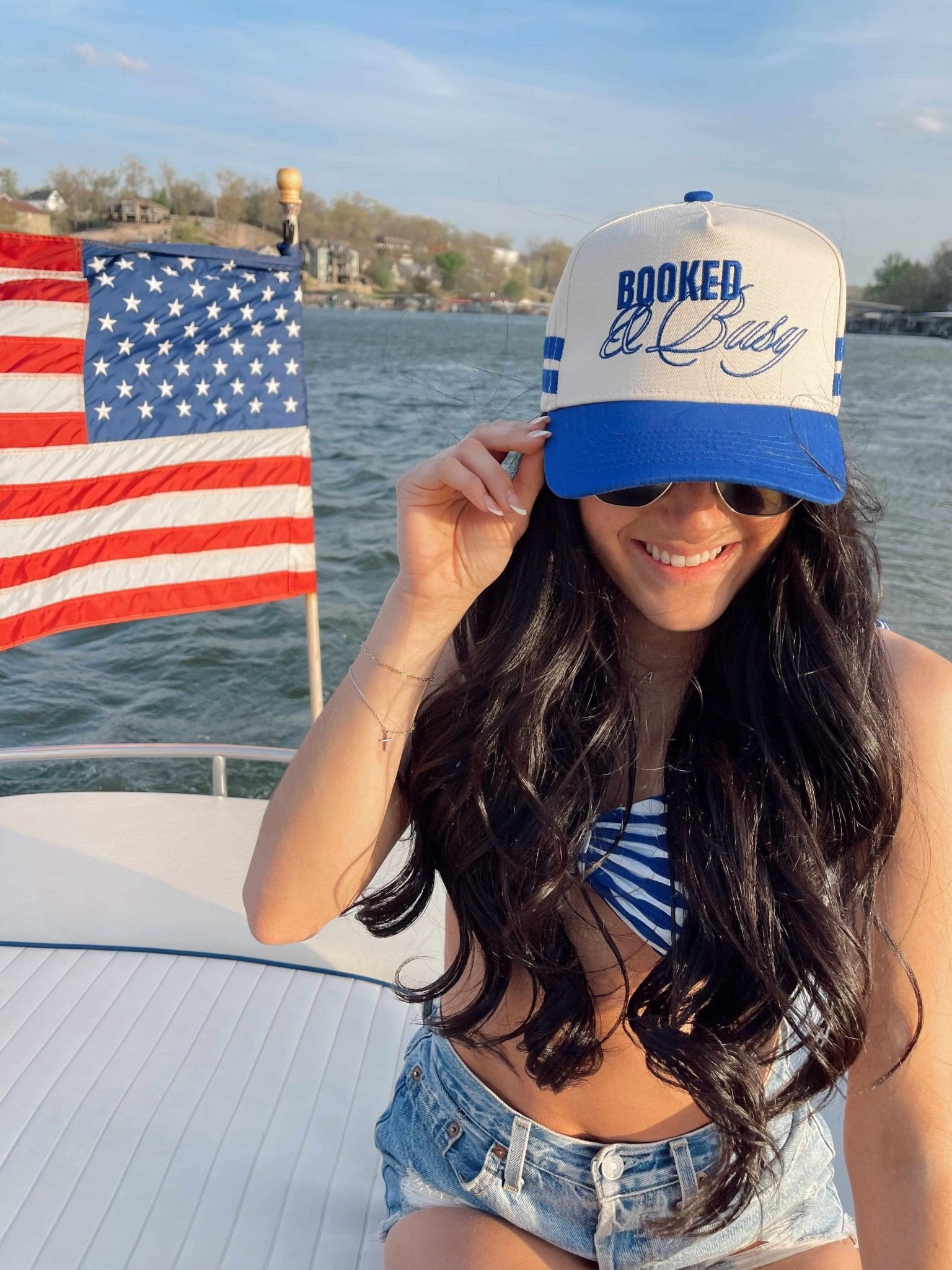 White and royal blue trucker hat with "Booked and Busy" text, worn on a boat with American flag in background